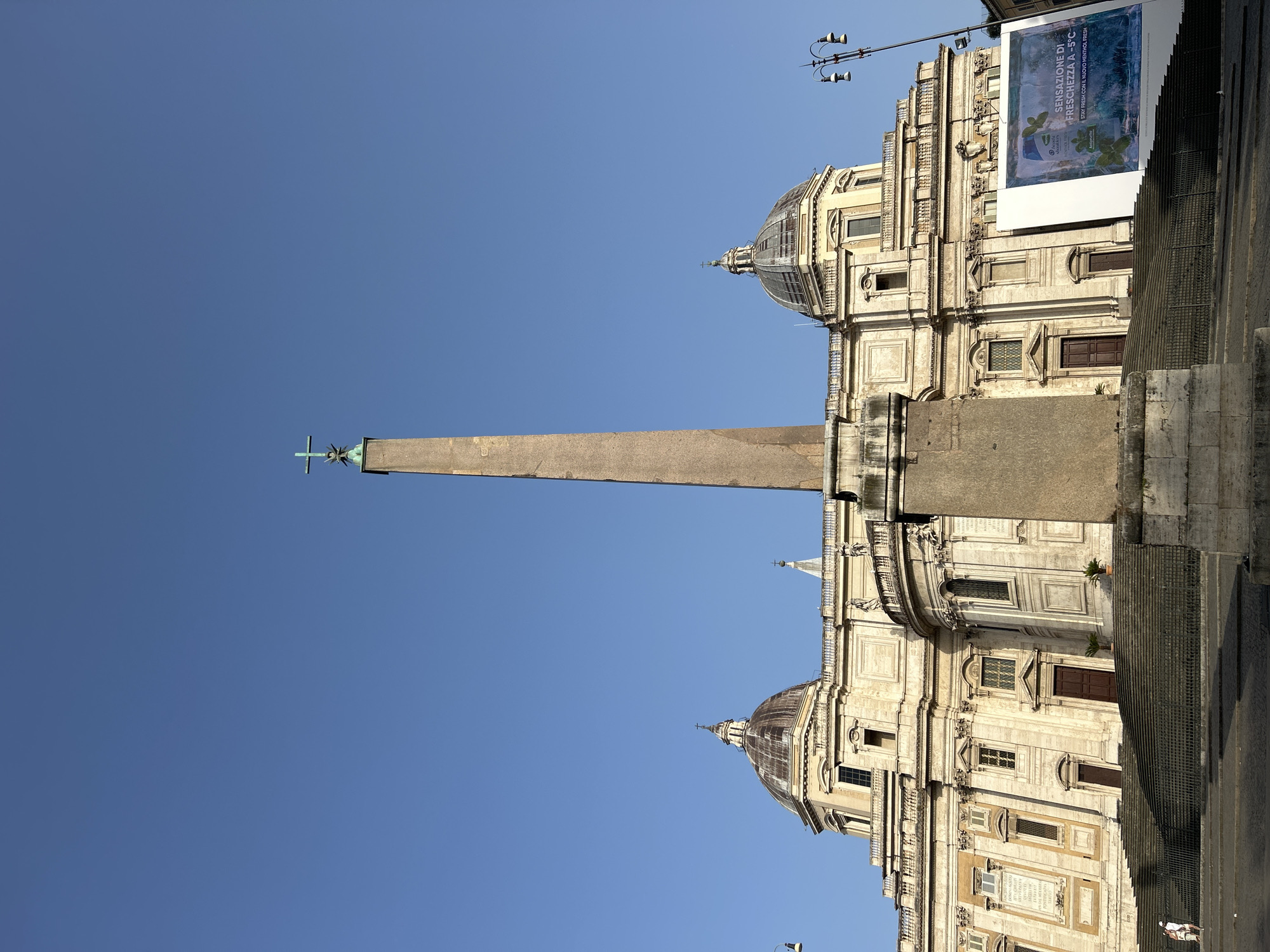 The image shows the Basilica di Santa Maria Maggiore in Rome, Italy. The front facade of the basilica is visible, with its prominent staircase and twin domes. In front of the basilica stands a tall Egyptian obelisk topped with a cross. The sky is clear and blue. A large banner with an advertisement is displayed to the right of the basilica. There are a few people walking on the steps and in the square in front of the basilica. The scene is illuminated by sunlight, suggesting it is daytime.
