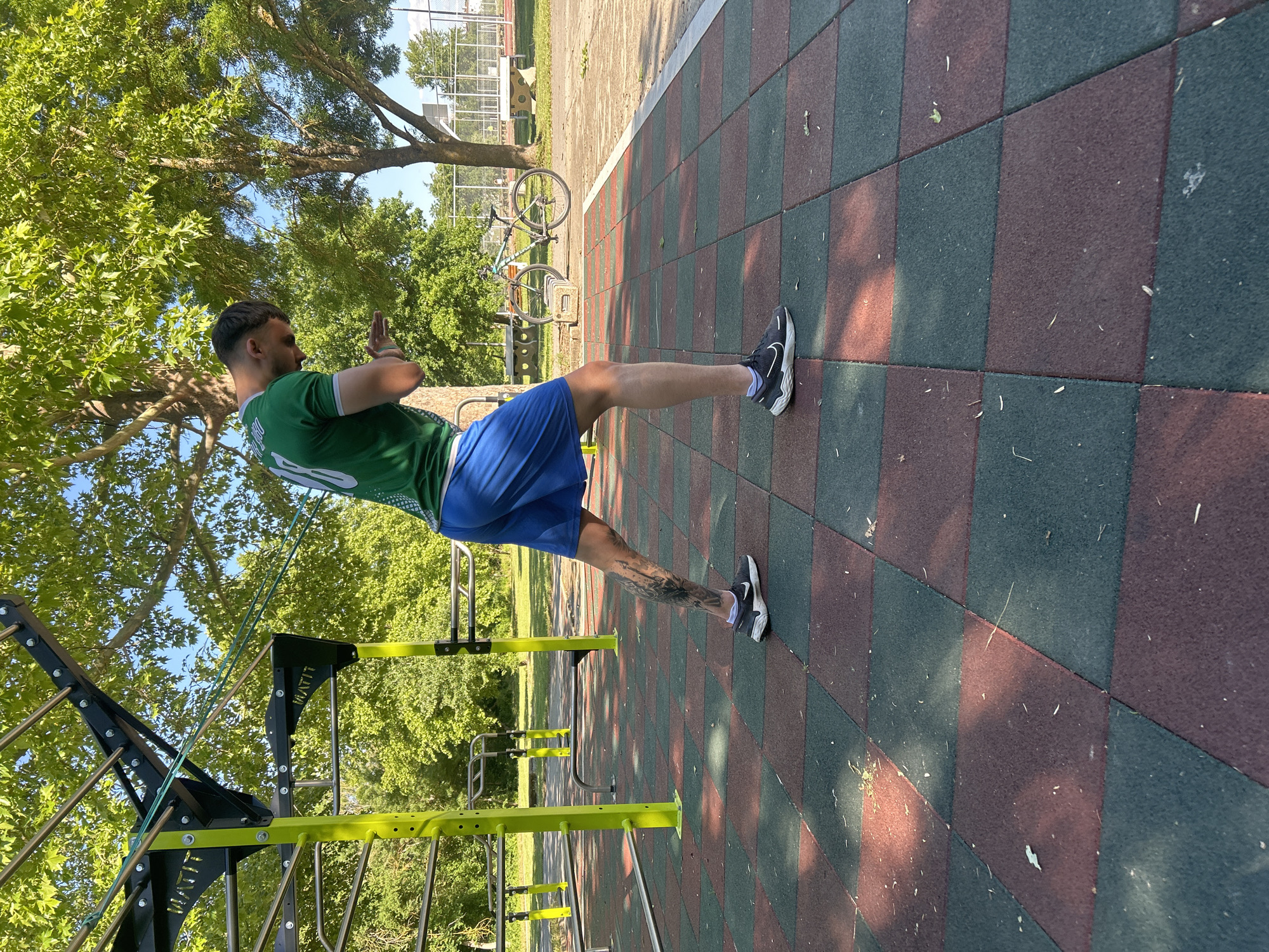 A man is exercising outdoors in a park-like setting. He is positioned between an outdoor exercise structure and a tiled surface, possibly in Elhovo, Bulgaria. The man is wearing a green shirt with white markings, blue shorts, and black and white athletic shoes. He has tattoos on his lower leg. His hands are clasped together in front of him at chest level. The exercise structure is black and yellow, featuring a ladder-like apparatus. The ground is covered in alternating green and red-brown tiles. In the background are trees, another exercise apparatus, and a bicycle rack. The scene is lit by sunlight, casting shadows on the ground.