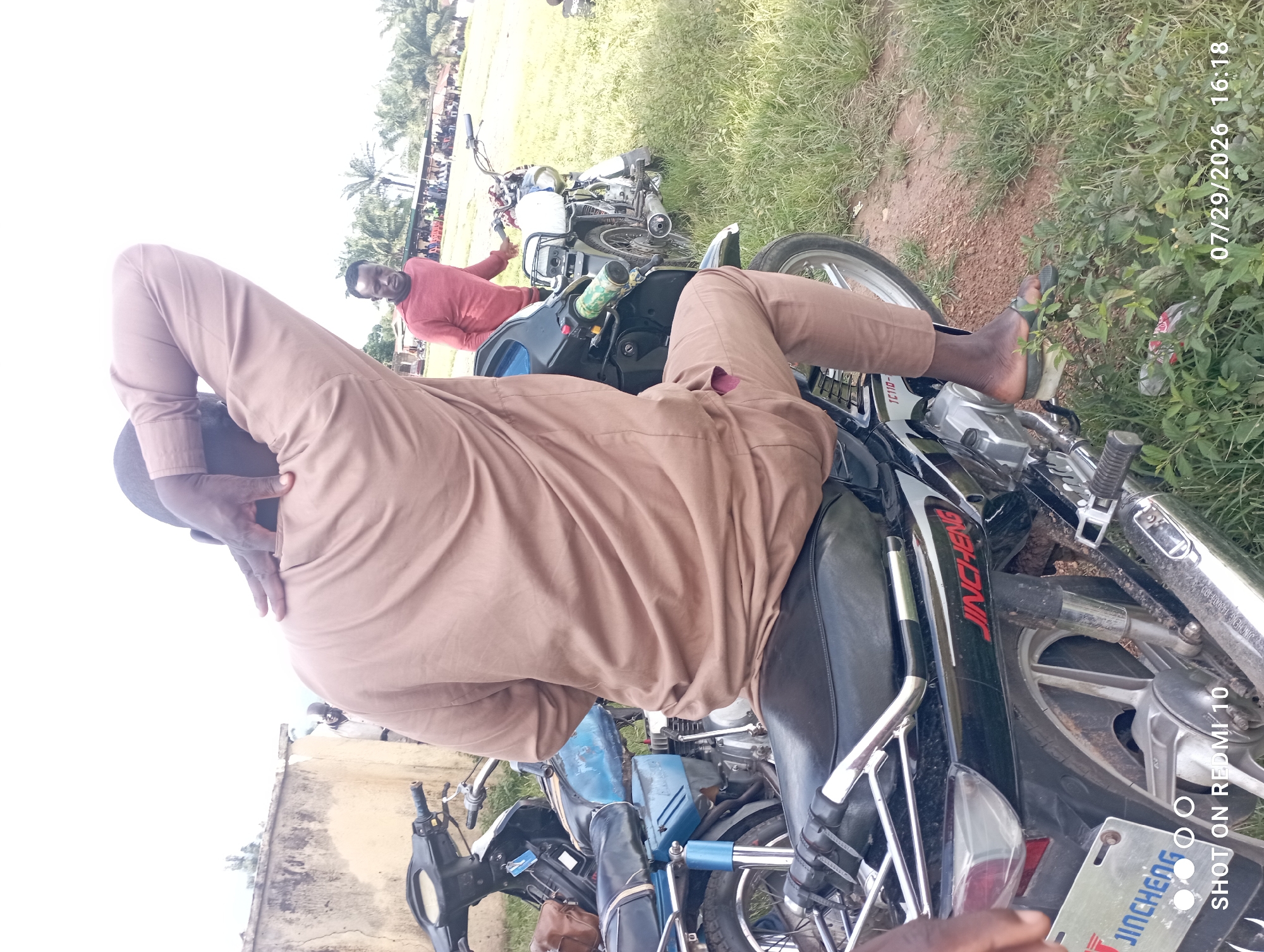 The image shows a man sitting on a black motorcycle in Serti, Nigeria. He is wearing a tan long-sleeved shirt and pants. His right arm is raised behind his head. Another man in a red shirt is standing behind him, looking towards the camera. In the background, there are more people and palm trees. The scene appears to be outdoors in a grassy area, with a building wall in the far left background. The motorcycle has a blue fuel tank and a silver exhaust pipe. The timestamp in the lower right corner indicates July 29, 2026, at 16:18. The text "SHOT ON REDMI 10" is visible in the lower left corner, along with a license plate with "JINCHENG" written on it.
