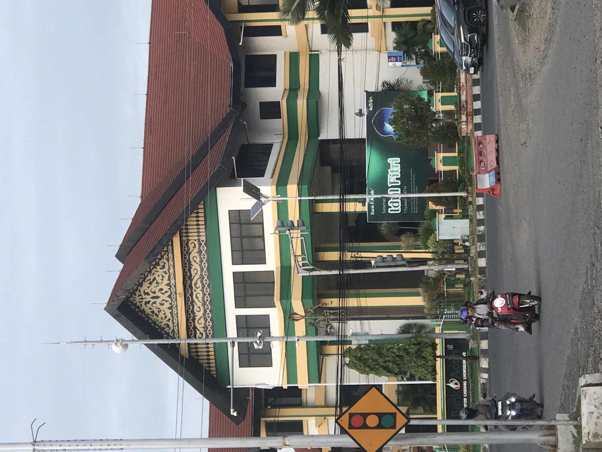 The image captures a street scene in Lhokseumawe, Indonesia, featuring a distinctive building with a red tile roof and a façade adorned with a green and yellow striped design and traditional Aceh patterns. A large banner advertising "Selamat Idul Fitri" for the Bank Aceh is prominently displayed on the building.
The street in front of the building shows some motorcycle traffic, including one carrying multiple people. A utility pole displaying a diamond-shaped traffic signal warning of traffic lights ahead is located near the viewer. A parked black car is visible on the street.
The environment appears to be urban, with a cloudy sky in the background. The scene suggests a bustling city street with a blend of local architectural style and contemporary transportation.