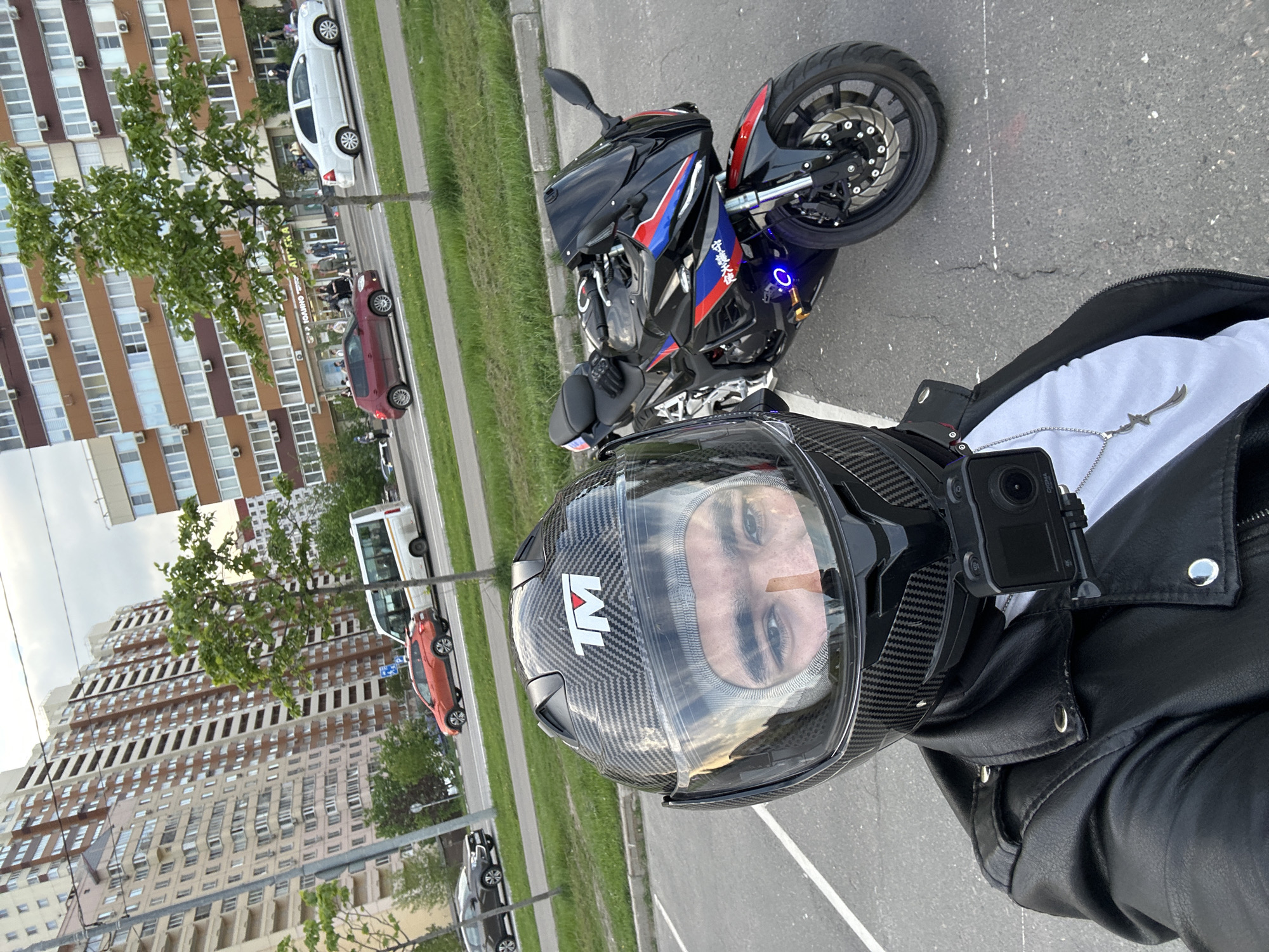 A first-person perspective photograph shows a person wearing a carbon-fiber motorcycle helmet with a transparent visor, through which their face is visible. A black action camera is mounted on the left side of the helmet. The person wears a black leather jacket, with a white shirt underneath, and a silver chain with a pendant is visible. A black and blue sport motorcycle is situated behind the person, parked on an asphalt surface. In the background, a row of cars are parked alongside a grassy area. Tall apartment buildings are visible behind the cars and trees. Based on the architecture of the buildings, the image likely shows a location in Moscow, Russia.