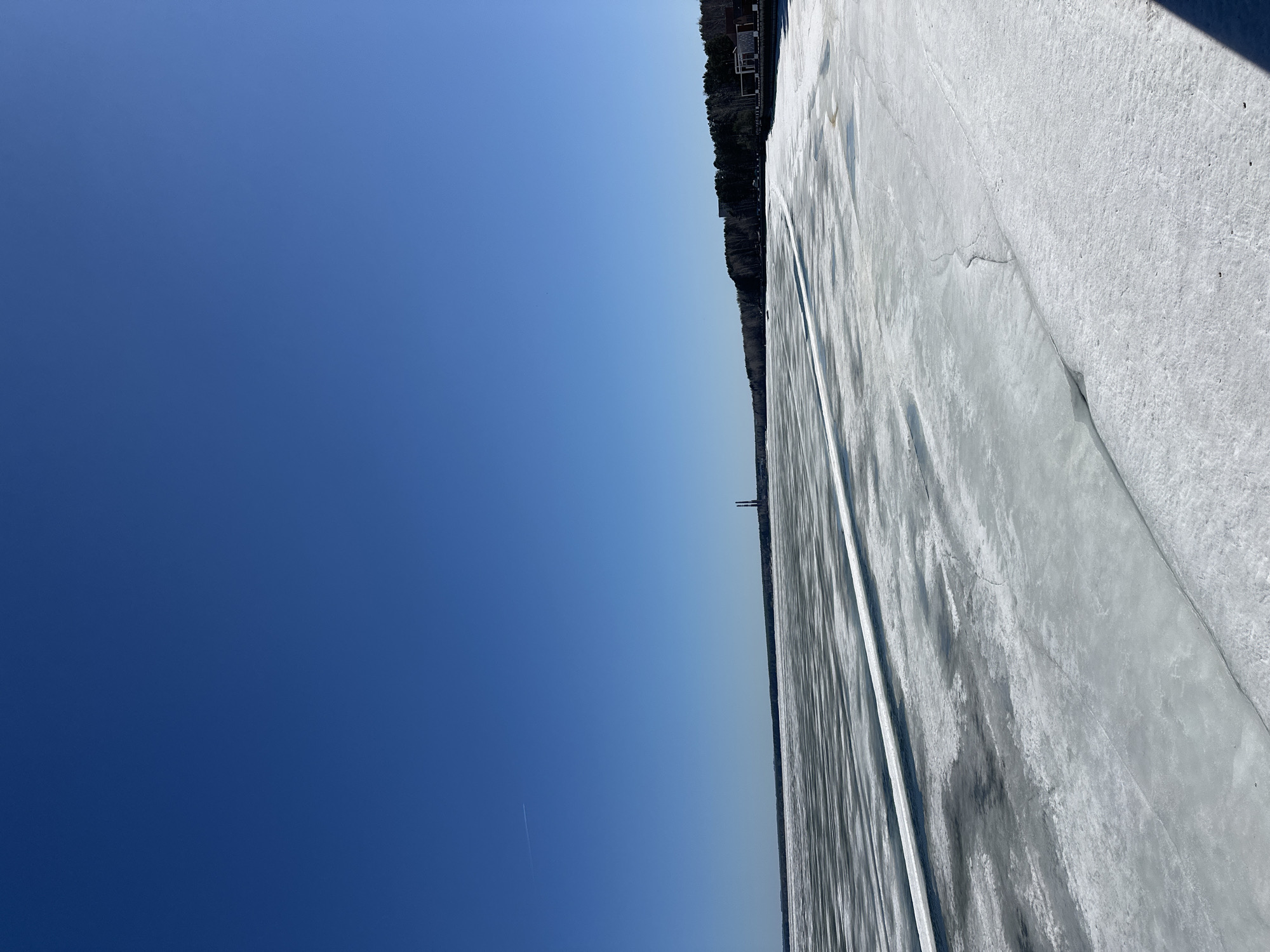 The image shows a frozen body of water, likely the Kama Reservoir, adjacent to the city of Chaikovsky, Russia. The ice covers most of the lower right half of the frame, exhibiting patterns and cracks. Along the shoreline, a line of trees and a few buildings are visible. The sky is clear and blue, occupying the upper left portion of the image. There is a contrail visible in the sky, suggesting an airplane is nearby.
