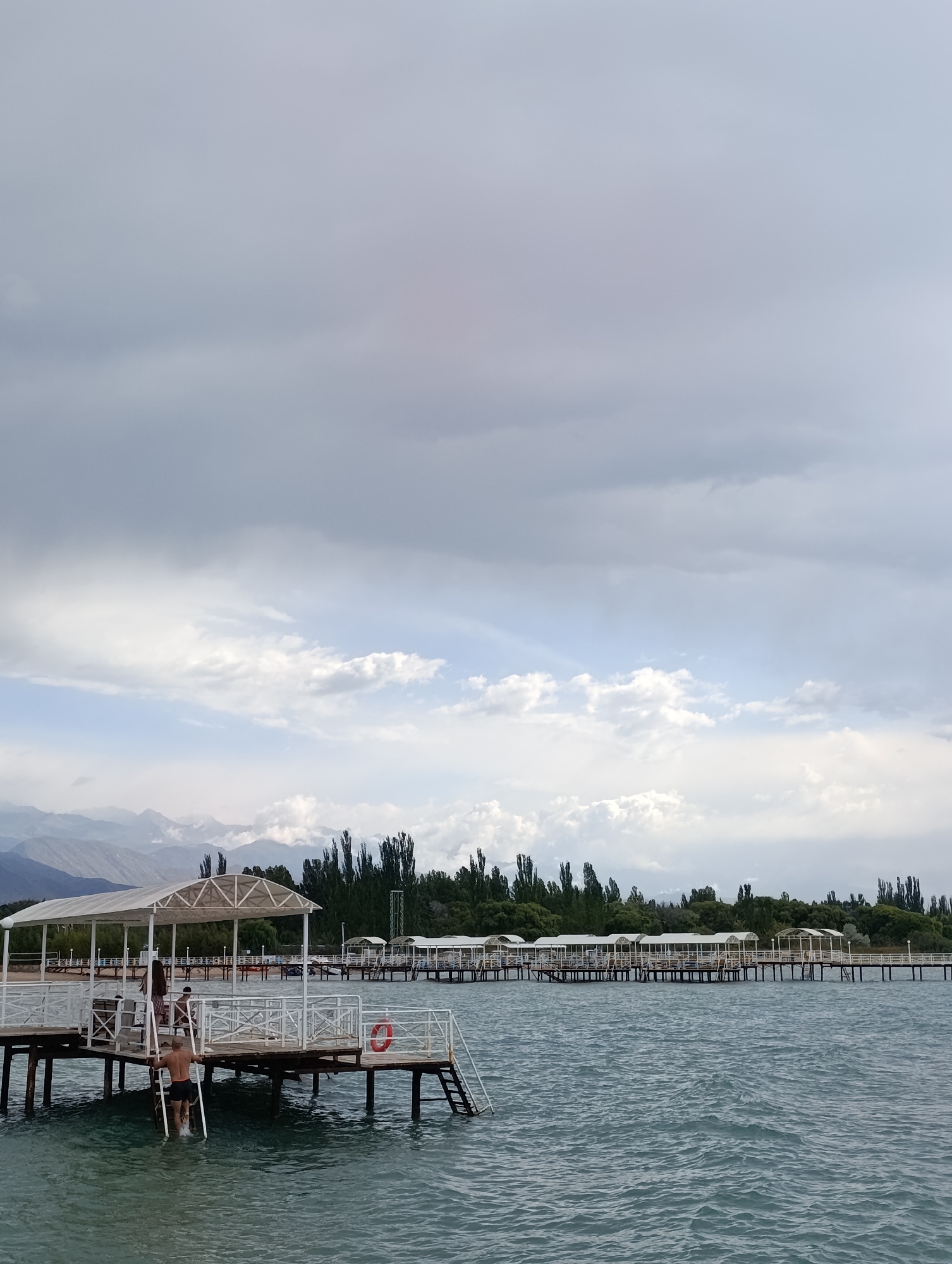 The image depicts a scene in Sary-Oy, Kyrgyzstan, featuring a pier extending into a body of water under a cloudy sky. The pier is constructed with wooden planks and supported by dark vertical posts. It is lined with white railings and features covered sections with white roofs, resembling cabanas.
On the pier, several individuals are visible. One man, wearing black shorts, is climbing up a ladder from the water onto the pier. Two women, one in a patterned dress and another with long dark hair, are standing further back on the pier. A child is also present near the ladder.
In the distance, across the water, is a shoreline with a dense line of tall, slender trees. Behind the trees, a range of mountains with a misty, layered appearance is visible against the sky. The water is a deep blue-green, with visible ripples and waves. A red life preserver is mounted on the railing of the pier. The overall atmosphere is overcast, with the sky dominated by shades of grey and white clouds.