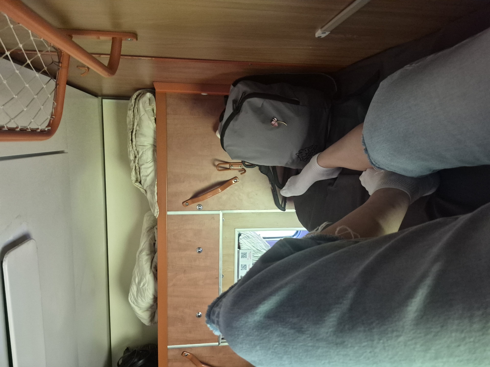 The image depicts a close-up, low-angle view of two pairs of legs, one draped over the other, inside a train compartment. The legs are clad in blue denim jeans and white socks. The compartment features wooden paneling, shelving with folded blankets, and a grey backpack with a floral pin. A metal rack and hooks are visible on the right wall. The lighting is artificial and appears to be from within the compartment. The scene suggests a moment of rest or travel within a train.