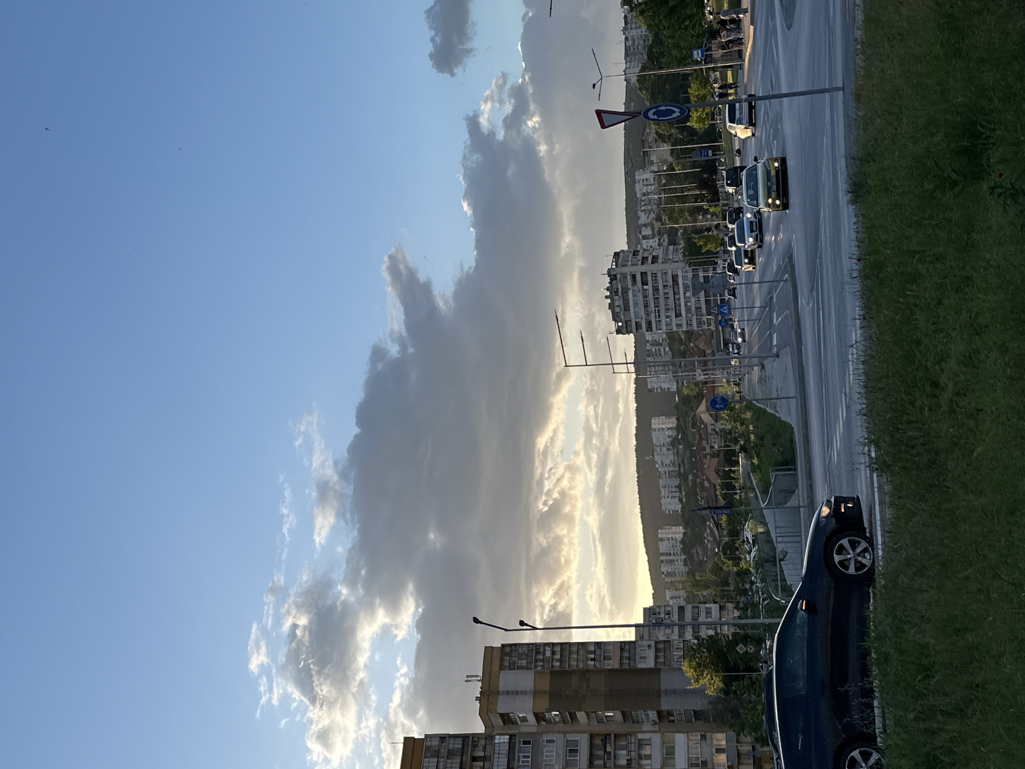 The image depicts a city street with several buildings and cars. The sky is visible with a mix of clear blue and thick, dark clouds. Along the right side of the image, a road is visible with several cars driving in a line. A narrow patch of green grass is adjacent to the road. On the opposite side of the road, numerous apartment buildings are seen, gradually ascending towards a hill in the background. Streetlights and traffic signs are interspersed along the road. A dark-colored car is parked on the right edge of the foreground.