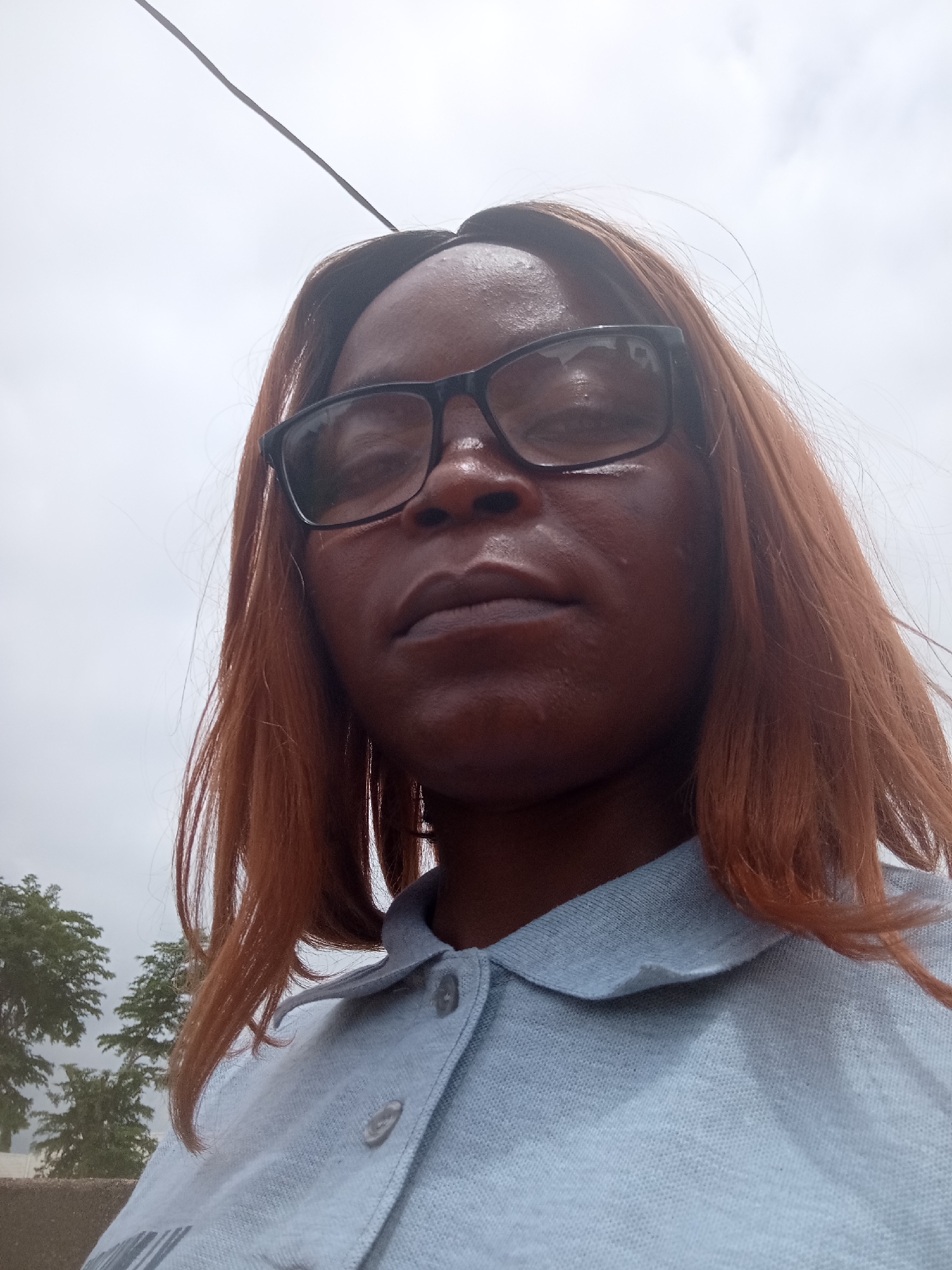 The image is a close-up, low-angle selfie of a Black woman with copper-colored hair, wearing glasses and a light blue polo shirt. She is outdoors, with a cloudy sky in the background and some green trees visible on the left. A black wire is also visible extending from the top left corner of the frame. The woman's face is the primary focus, with her mouth slightly open and her eyes looking towards the camera. The overall impression is of a candid moment captured outdoors, potentially on a slightly overcast day. The specific location within Kason, Nigeria, is not discernible from the visual information alone.