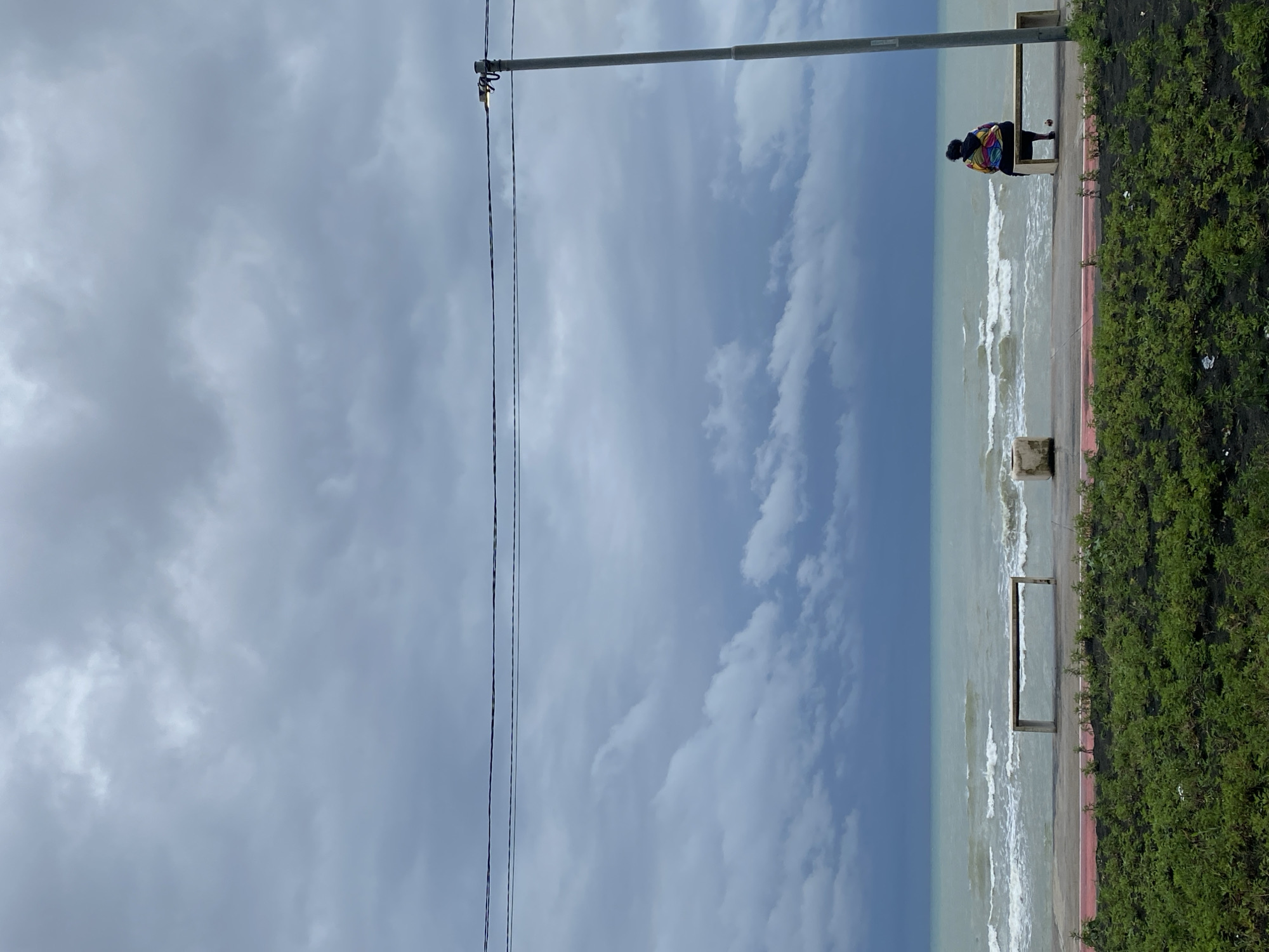 The image depicts a cloudy day at the Black Sea coast in Ureki, Georgia. In the foreground, lush green bushes are visible. Beyond the bushes is a paved walkway with a red stripe. A concrete bench sits on the walkway, empty. Further back, a person wearing a colorful hooded jacket is seated on another concrete bench, facing the sea. The sea itself is a light turquoise color with moderate waves. The sky is filled with heavy, dark grey clouds. A tall, gray lamppost stands on the right side of the image, with wires running horizontally across the frame.