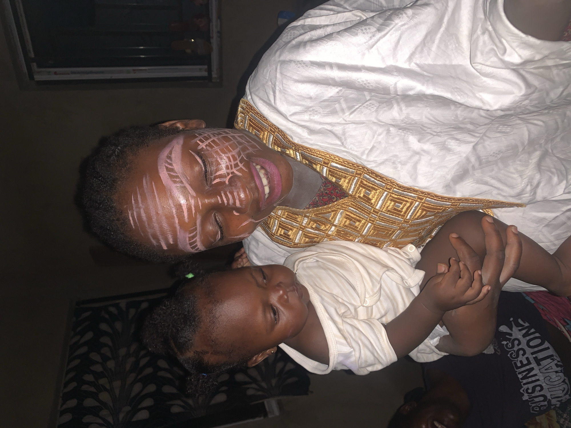 In Agban, Nigeria, an adult is holding a baby. The adult has white and pink face paint on. The adult is wearing a white garment with gold trim. The baby is wearing a light-colored onesie. In the background, there is a dark curtain with a pattern and a window. Another person is visible in the lower left of the image, wearing a black shirt with white text. The lighting suggests the photo was taken indoors at night.