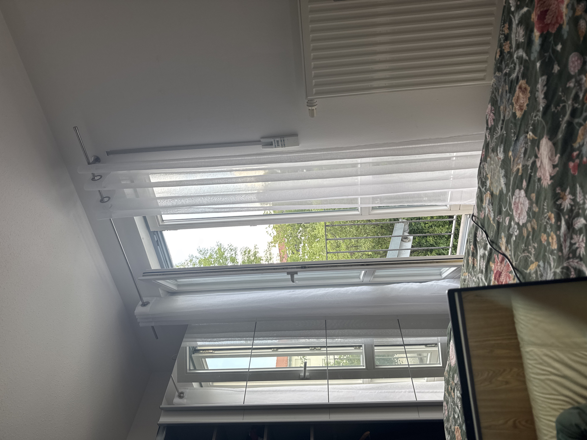 This media file depicts an interior scene looking upwards towards a slanted ceiling with a skylight. The main subjects are architectural elements and household furnishings. A prominent feature is a large, multi-paned skylight that is partially open, allowing a view of green trees and a glimpse of buildings and a metal railing outdoors. Beneath the skylight, sheer white curtains are hung on a track system, creating a layered effect. A metal radiator is visible mounted on the ceiling above the curtains.
On the right side of the frame, a section of a bed with a floral-patterned duvet cover is visible. The duvet features a dark green base with a dense pattern of flowers in shades of pink, red, yellow, and blue. A black cable runs across the bedspread. A laptop or similar electronic device with a black bezel is positioned in the lower right corner, reflecting the light from the window.
The scene appears to be in a residential setting, likely a bedroom or living area with a sloped ceiling, possibly an attic conversion. The presence of the bed and the soft, natural light suggest it is daytime, possibly morning or afternoon, with no indication of specific weather conditions beyond general daylight. There are no people visible in the frame.
Notable details include the two-tiered curtain system, the opening mechanism of the skylight, and the detailed floral pattern on the bedding. The overall impression is one of a domestic interior bathed in natural light. No visible text is present in the image. The location context provided is Höhenkirchen-Siegertsbrunn, Germany.