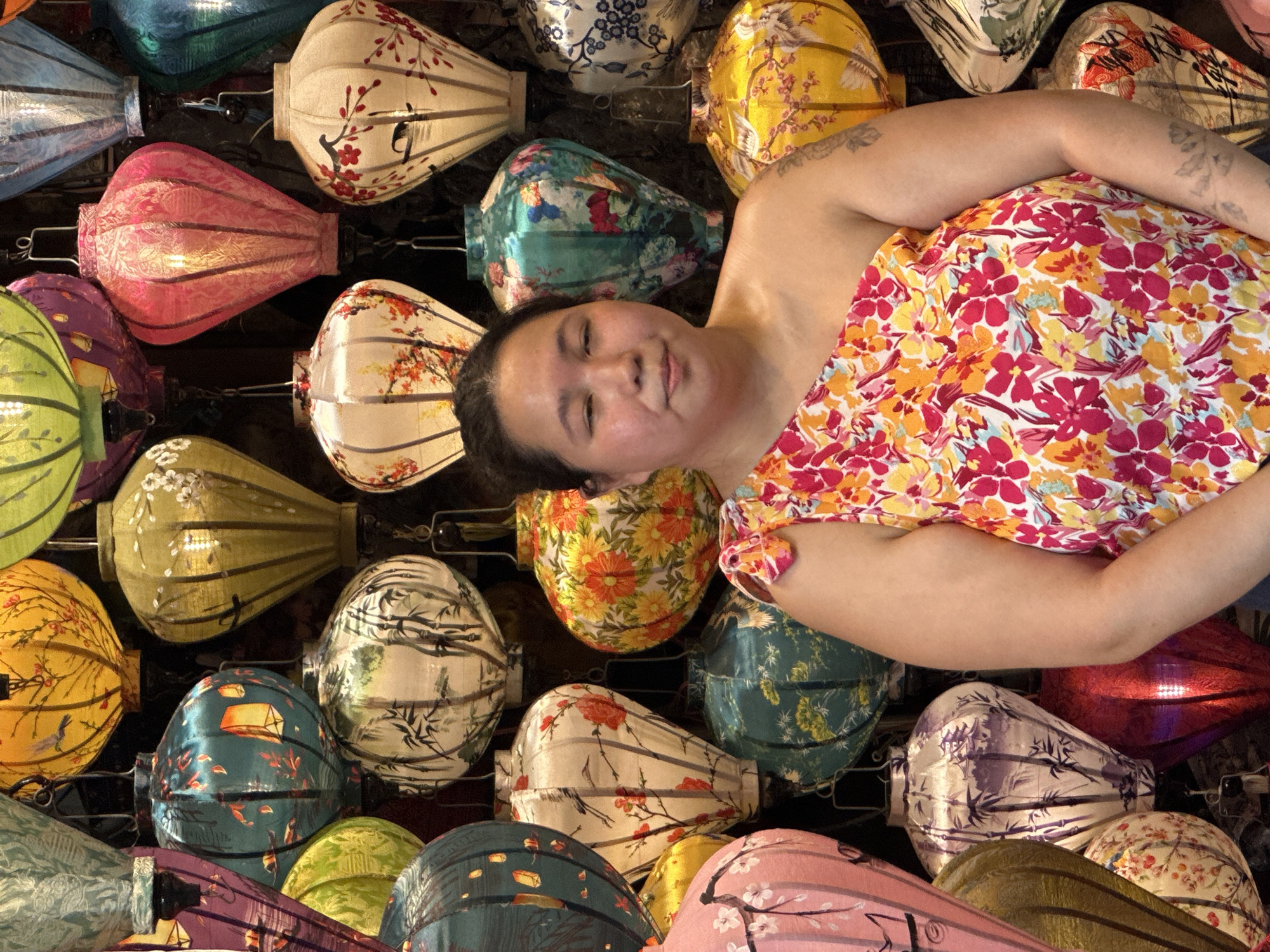 This media file features a person lying amongst a collection of decorative lanterns. The person, positioned in the center of the frame, is wearing a vibrantly patterned shirt with floral designs in pink, red, orange, and yellow. Their face is turned towards the camera, with closed eyes and a relaxed expression, suggesting a moment of rest or sleep. Their arms are resting on their body, partially obscuring their floral garment.
The background is densely populated with numerous paper lanterns of various shapes, sizes, and colors, all illuminated from within, creating a warm and inviting ambiance. The lanterns display a diverse range of artistic motifs, including floral patterns, cherry blossoms, birds, bamboo, and abstract designs. The color palette of the lanterns ranges from soft pastels like cream and pink to richer hues of green, yellow, blue, teal, and red.
The scene appears to be an indoor setting, possibly a shop or a market stall in Hội An Ward, Vietnam, given the prevalence of traditional Vietnamese lanterns. The arrangement of the lanterns suggests they are for sale or display. The lighting from the lanterns creates a soft glow, and the overall mood is tranquil and visually rich. There is no visible text in the media file.