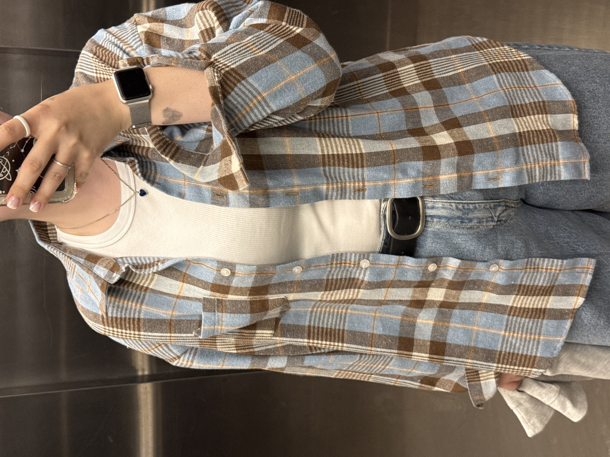 A person is standing in an elevator, facing the camera. They are wearing a blue and brown plaid button-up shirt over a white ribbed tank top, light-wash jeans, and a black belt with a silver buckle. A silver smartwatch is visible on their left wrist. Their right hand is holding a smartphone, and they are wearing rings on multiple fingers. The elevator walls are a dark, reflective metallic material. The scene is indoors, in what appears to be a standard elevator. The location is not specified within the image.