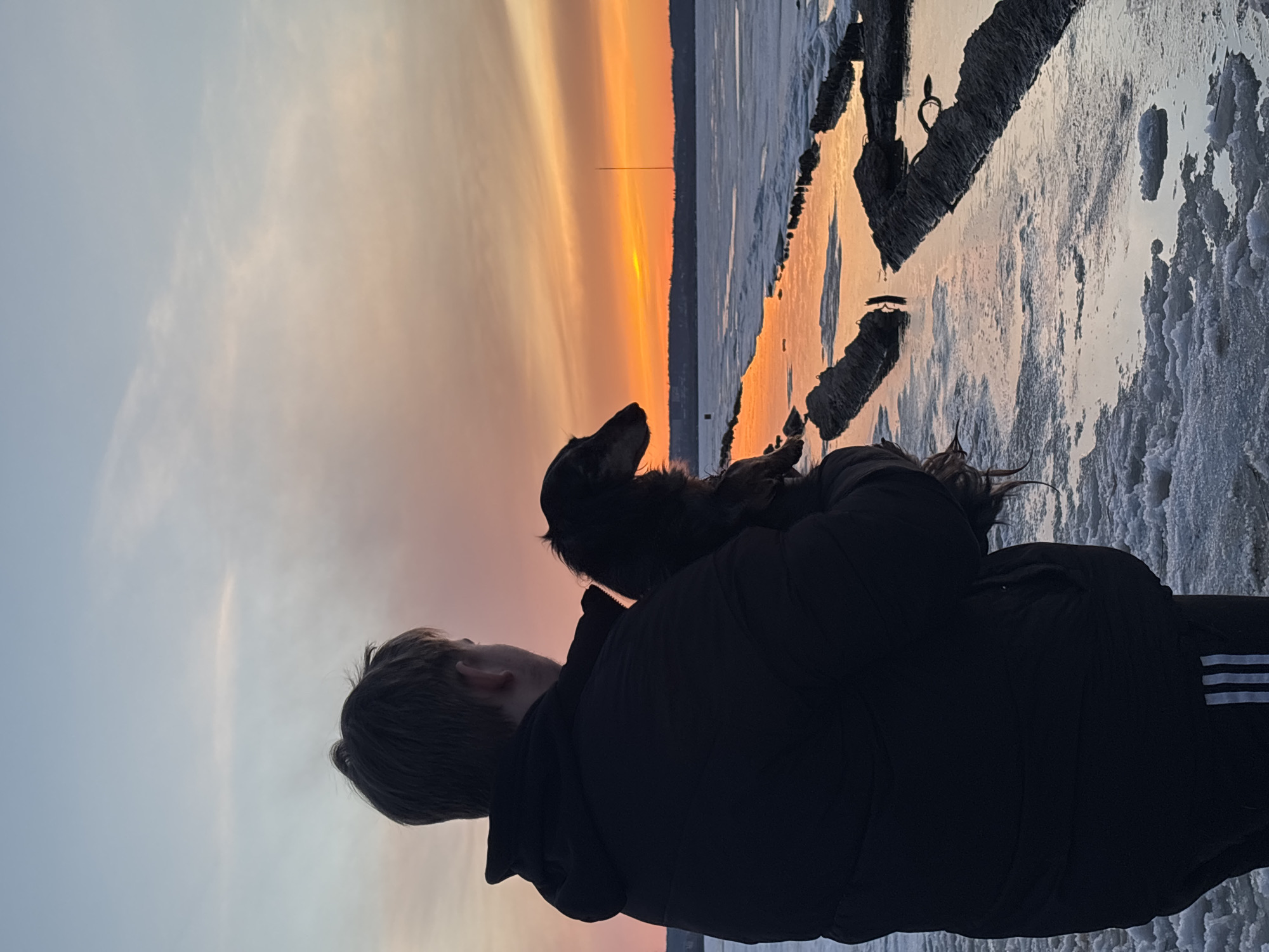 A person wearing a dark jacket with Adidas stripes on the sleeve is holding a dog, possibly a dachshund, against a sunset backdrop. The scene appears to be located near a body of water, potentially the Izhevsk Pond in Izhevsk, Russia, based on the skyline and surrounding environment. The area is partially covered with ice and snow. A tall structure is visible in the distance against the colorful sky. The person and dog are silhouetted against the bright sunset.