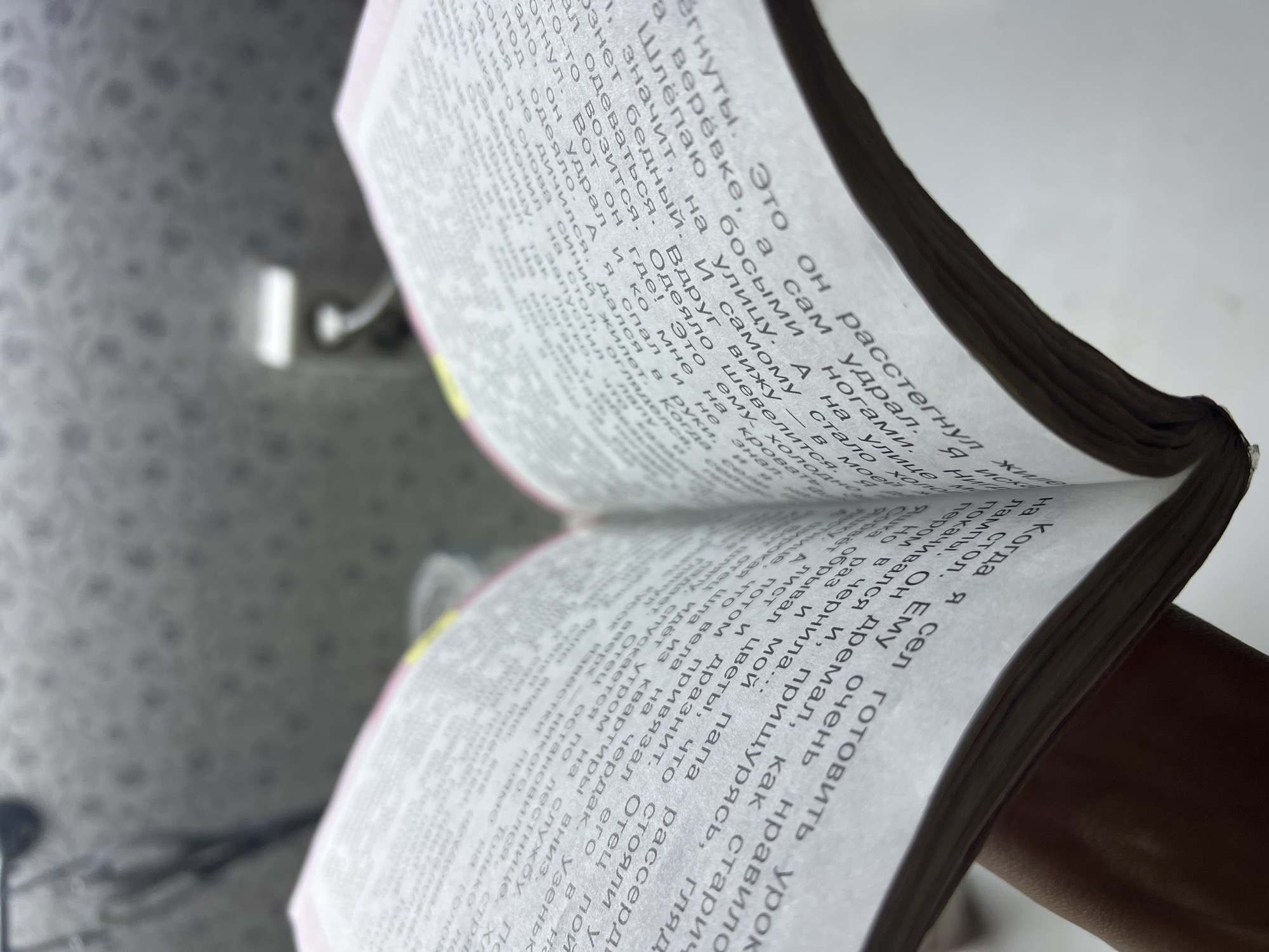 The image depicts an open book with Russian text printed on its pages. The book is held, suggesting someone is reading or displaying it. The text is dense, resembling literature or academic material. Due to the angle and limited clarity, the finer details of the environment are not discernible. It is difficult to provide information regarding the city of Noviy, Russia, due to limited visible content.