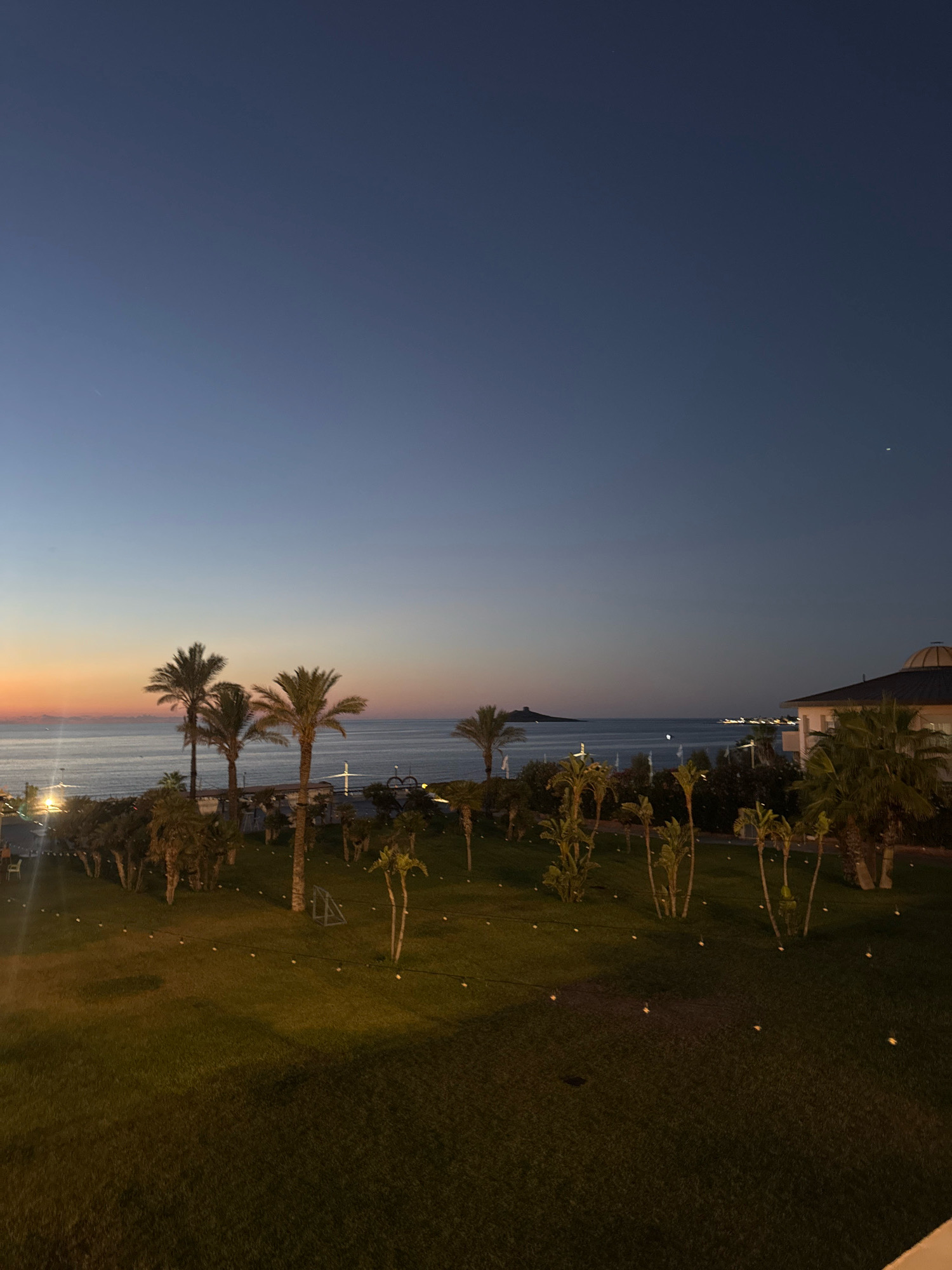 The image depicts a twilight scene overlooking the sea. The sky is a deep gradient of blue, transitioning to orange and pink hues near the horizon, suggesting either sunrise or sunset. Several palm trees are scattered across a well-maintained grassy lawn in the foreground. In the middle ground, the sea stretches out, with a few distant lights visible along the coastline. On the right side of the image, a building with a domed roof is partially visible, indicating a resort or hotel. The overall atmosphere is serene and tranquil. The location is the city of Isola delle Femmine, Italy.