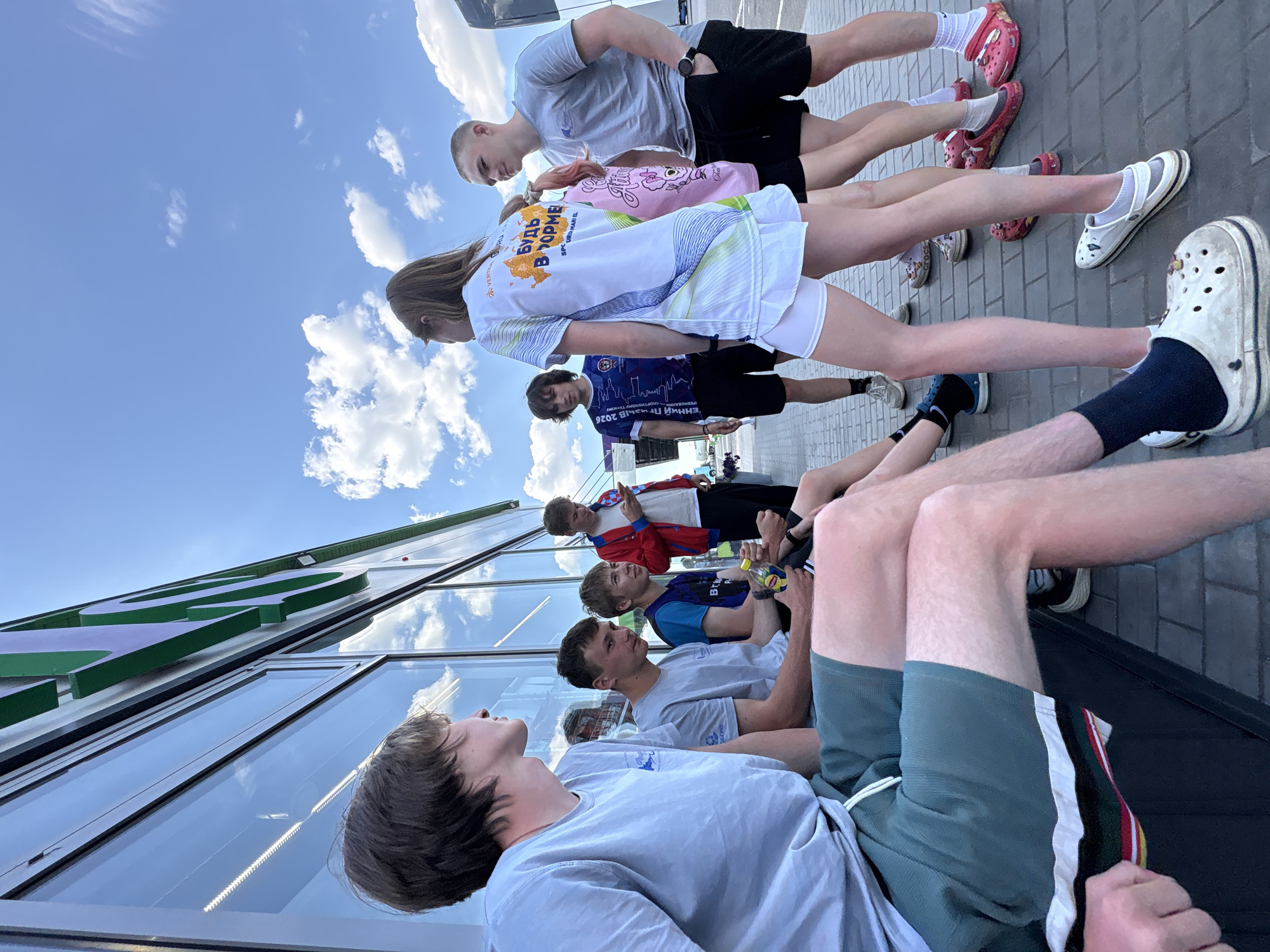 A group of at least 10 people, seemingly teenagers, are arranged in two lines, one sitting along a building facade and the other standing behind them, under a partly cloudy sky. The people in the front row are mostly wearing light-colored t-shirts and shorts. Several of them appear to be looking upwards. The people in the back row are wearing athletic attire. The building facade has large glass windows and green lettering. The ground is paved with gray bricks. The city is Казань, Russia.