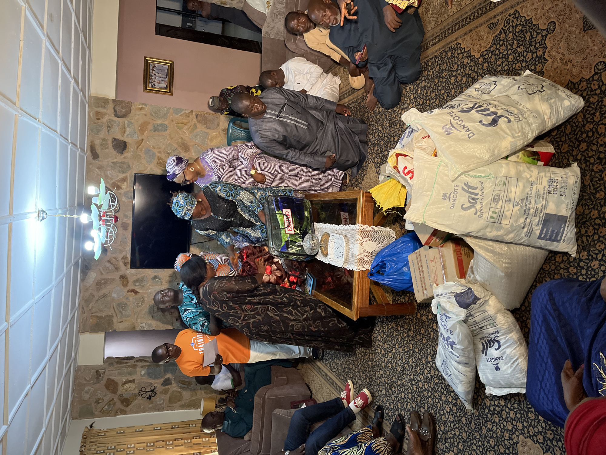 In Bukuru, Nigeria, a group of people are gathered indoors, likely in a living room. The room features stone-clad walls and a patterned carpet. A coffee table sits centrally, holding a crystal dish and a white lace runner. In the foreground, several large bags of "Dangote Salt" are prominent, along with other assorted bags and boxes.
The individuals in the scene are dressed in traditional African attire. Several men and women are interacting, with some appearing to be distributing or receiving items. One man in a grey agbada is kneeling, looking down, while another man in a dark blue outfit is seated and gesturing with his hand. A man in an orange t-shirt holds a piece of paper and appears to be addressing the group. The overall atmosphere suggests a distribution of goods or a community gathering.