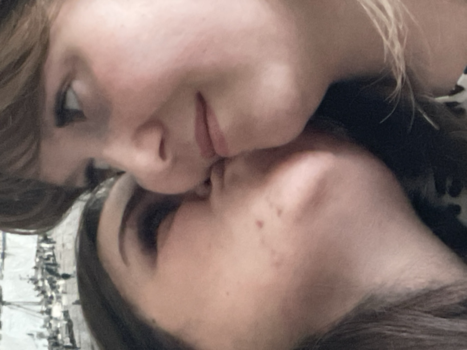 The scene captures two individuals in a close-up, head-and-shoulders frame. The individual on the left, presumed female with dark hair, has her face in profile, pressing her lips and cheek against the lower facial region of the individual on the right. Her visible eye is closed, and minor skin irregularities are present on her cheek. The individual on the right, presumed female with lighter hair, has her face angled slightly away from the camera, with her right eye open and directed upwards-right. Both individuals exhibit light skin complexions. The background is out of focus; a blurry, monochromatic image resembling a cityscape or port scene is partially visible in the upper left quadrant. No specific identifiers for a location within Chaikovsky, Russia, are present in the image.