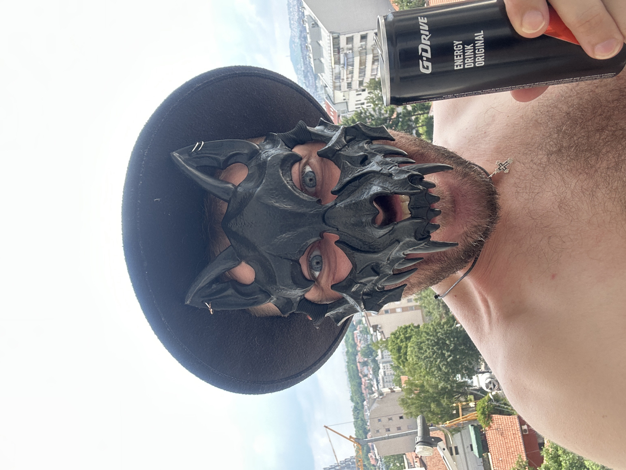 A man, shirtless and wearing a black wolf-like mask and a black hat, holds a can of G-Drive energy drink in his right hand. The man's face is partially obscured by the mask, but his blue eyes are visible. The background shows a cityscape with buildings, trees, and a cloudy sky, likely taken from an elevated position such as a balcony or rooftop in Zvezdara Urban Municipality, Serbia. A construction crane is visible in the middle ground to the left. The man has a cross necklace on his chest.