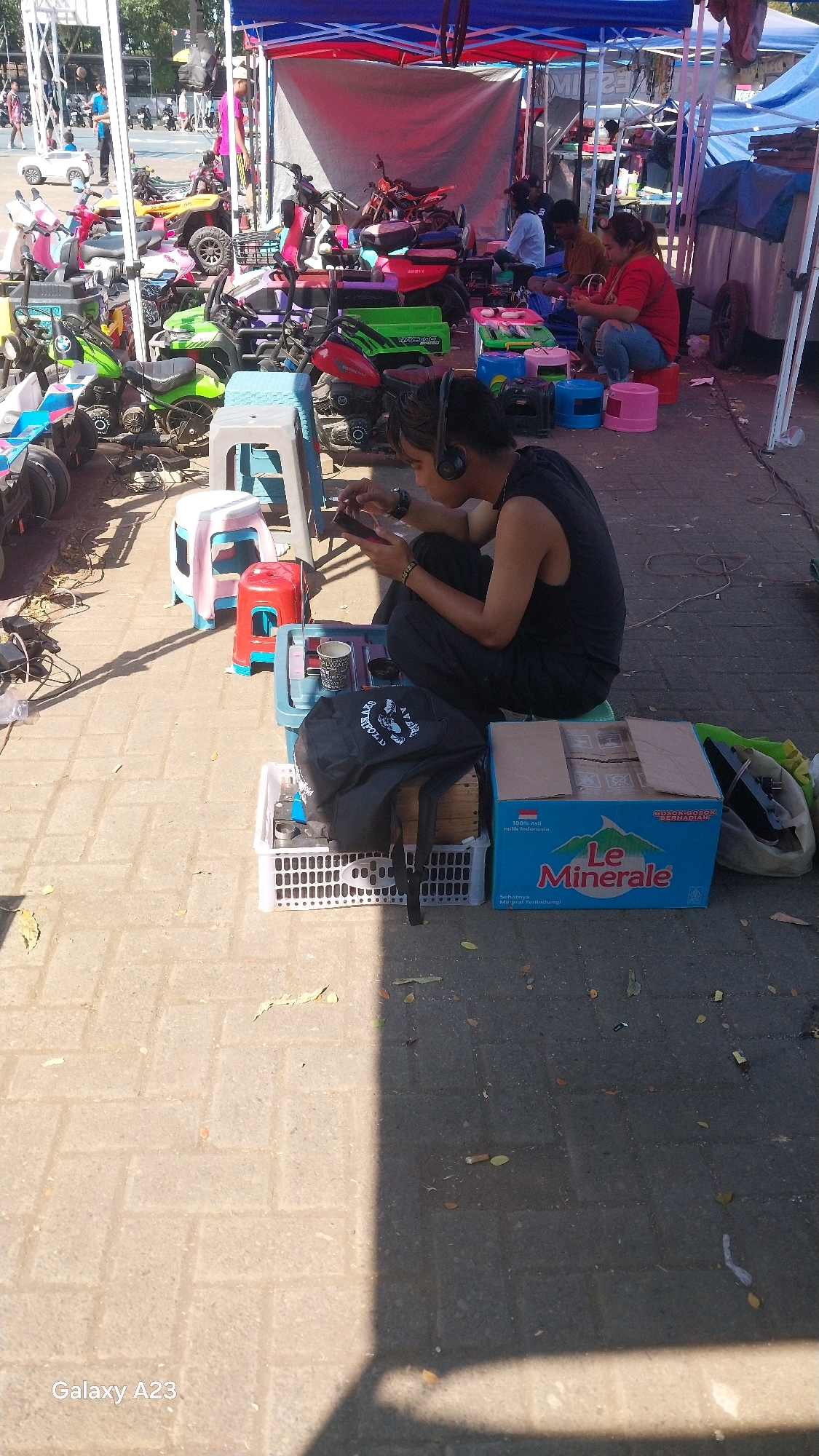 In Malang, Indonesia, a person wearing headphones and a black tank top is sitting on the ground, looking at their phone. They are surrounded by various items, including a black backpack, a blue and white crate, a red plastic stool, and a large blue box with the "Le Minerale" logo. In the background, several colorful children's electric vehicles are parked under a tent. Other individuals are visible in the distance, near more tents and stalls. The ground is paved with interlocking bricks, and shadows from the tents and structures fall across the scene. The overall impression is of an outdoor marketplace or event.