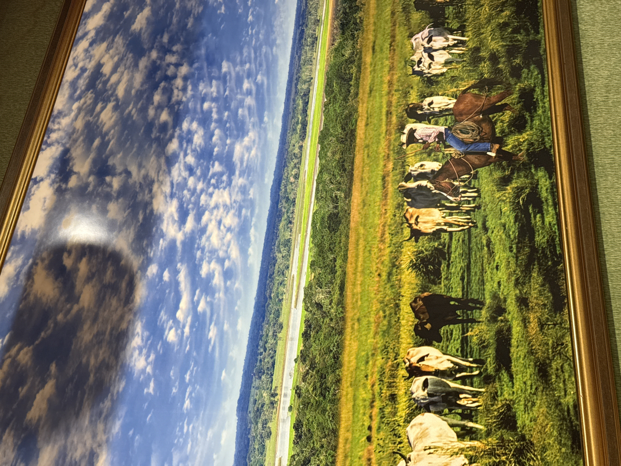 The image depicts a framed landscape painting. The scene shows a vast green field with a herd of cattle. A cowboy on horseback is visible in the foreground, guiding the cattle. In the background, a river winds through a densely forested area, with a range of hills or mountains in the distance. The sky is blue with scattered clouds. This image is located in the city of Torrejón de Ardoz, Spain.