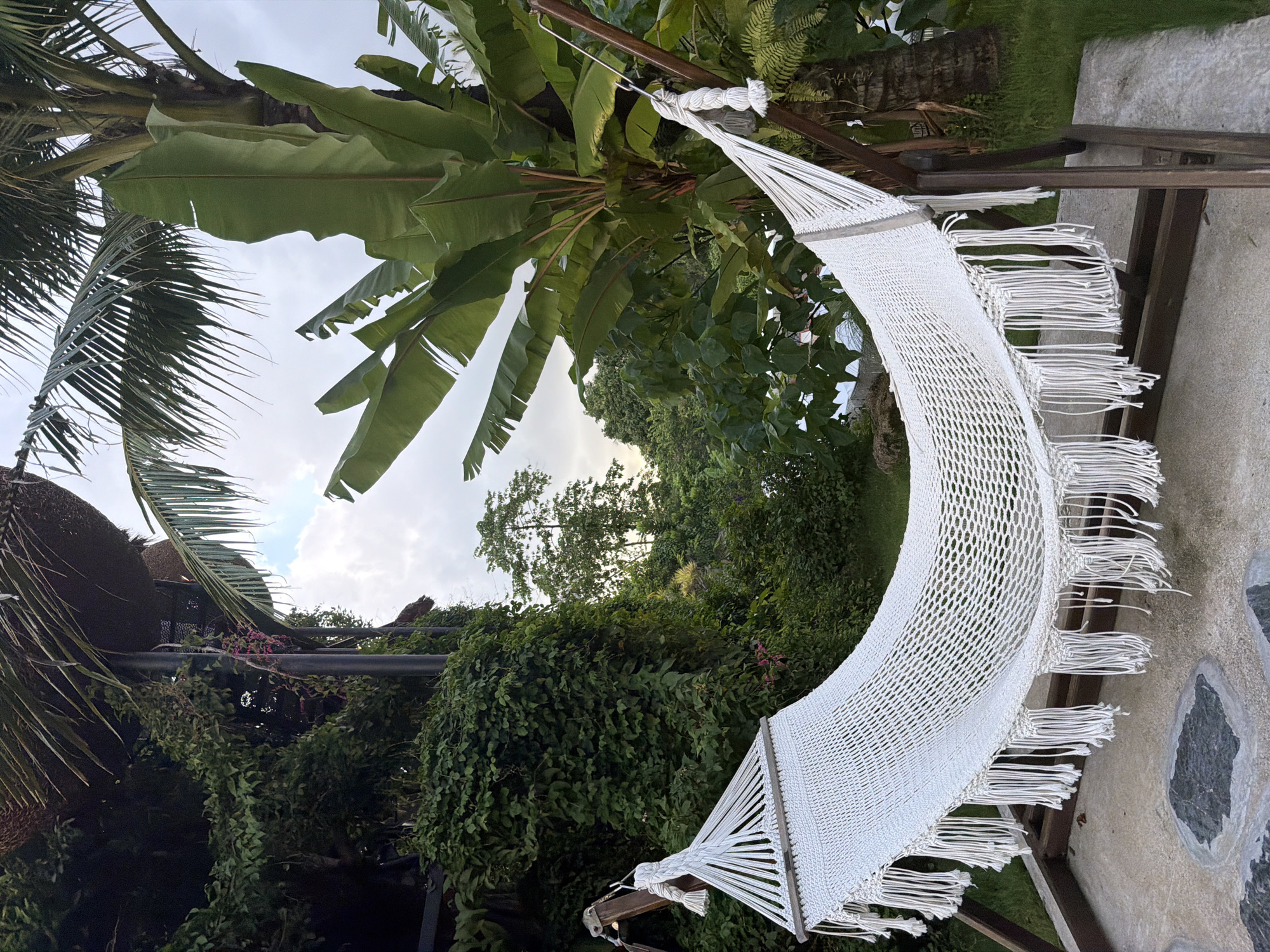 A white macrame hammock with fringe is suspended from a wooden stand in an outdoor setting. The hammock is positioned in the foreground, with lush green foliage and trees forming the background. To the left, a structure covered in ivy is visible, and a large banana tree with broad leaves is prominent on the right. The sky above is overcast with patches of light. The location appears to be a tropical or subtropical environment, consistent with Ban Khlong Khian, Thailand, suggesting a relaxing outdoor space.