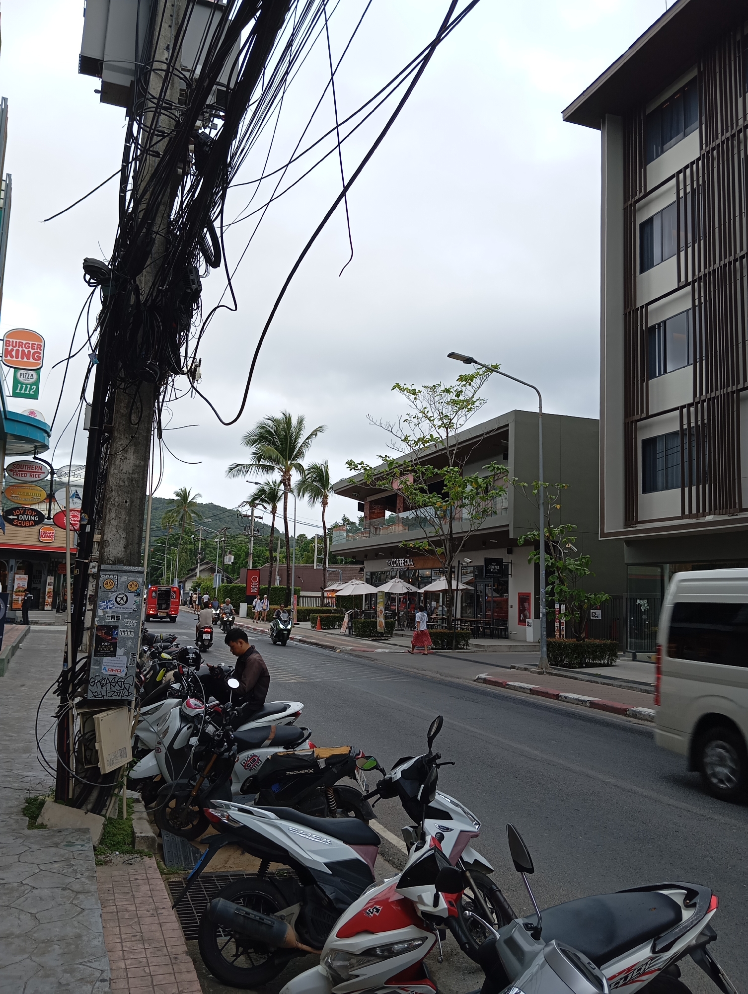 The image depicts a street scene in Karon, Thailand, characterized by a tangled mass of electrical wires overhead, a common sight in Southeast Asia. In the foreground, a row of parked scooters lines the sidewalk, some with visible branding like "ZOOMER-X." A man in a brown shirt is seated on one of the scooters. The sidewalk is paved with interlocking stones and bordered by a storm drain. To the left, a Burger King sign is visible, along with other signs indicating food and services. The street itself is paved and appears to be moderately busy, with several scooters and a red truck in motion. Palm trees are interspersed with buildings along the street. On the right side of the street, a modern building with louvred facade and multiple windows stands. The sky is overcast, suggesting a cloudy day. The overall impression is one of a bustling, somewhat cluttered urban environment.