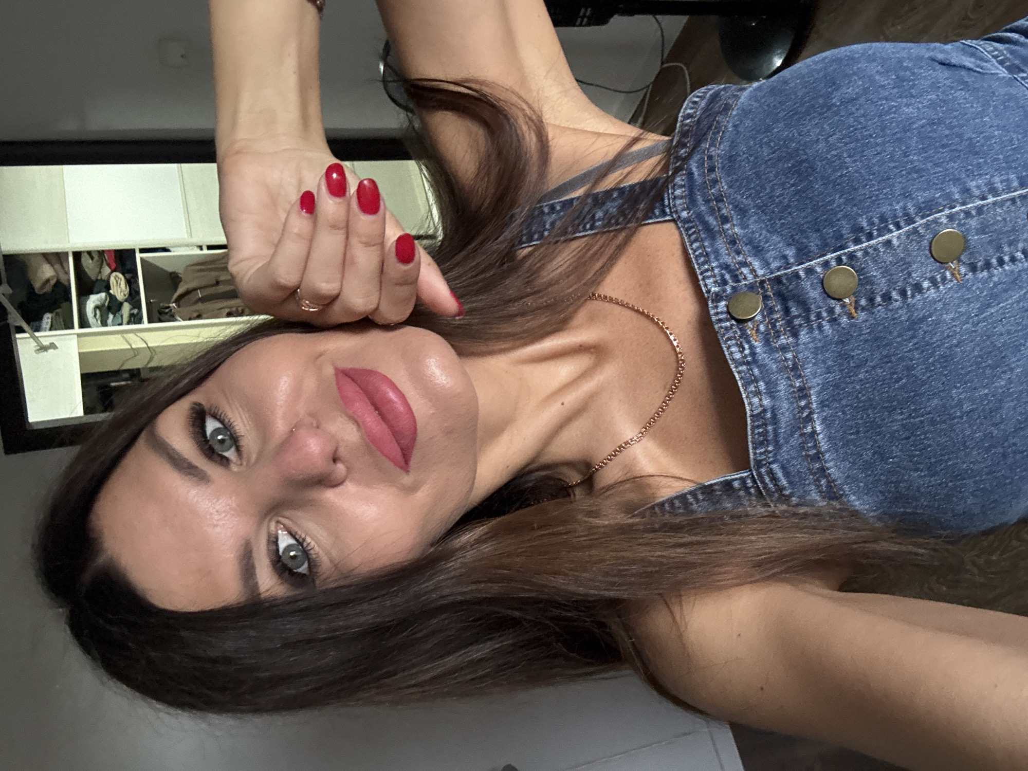 A selfie shows a woman with dark brown hair and green eyes, wearing a denim overalls dress. She is looking at the camera with her head tilted and her right hand raised to her cheek. Her nails are painted red. She has pink lipstick on and a gold necklace. In the background, a closet is visible with clothes hanging inside. The scene appears to be indoors, likely in a residential setting. There is no discernible information about the location in Chaikovsky, Russia, or any specific environmental details relevant to that city.