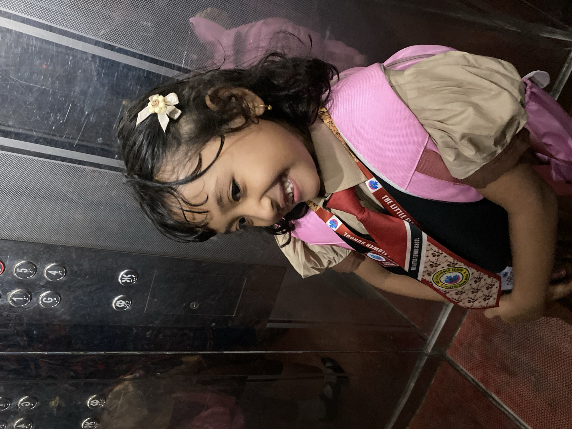 A young child is in an elevator. The child is wearing a school uniform, a pink backpack, and a hair clip with a flower on it. The uniform has "The Little Flower School" written on it. The elevator buttons are visible. The scene is possibly within Dhaka, Bangladesh, given the school name "The Little Flower School".