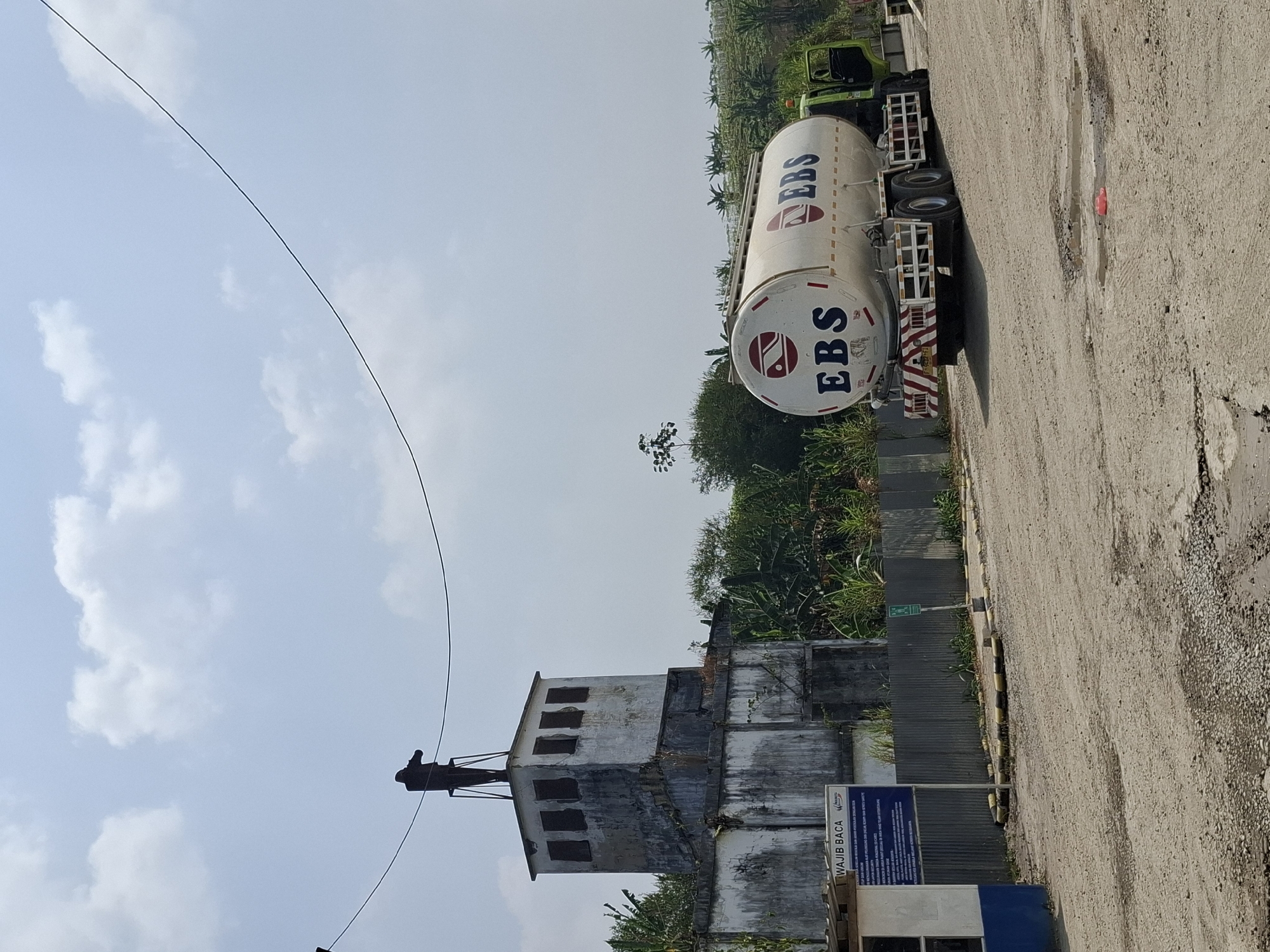 In Pamuruyan, Indonesia, a tanker truck with "EBS" branding is parked on a dirt road next to a dilapidated concrete structure. The structure has a tower-like top with a figure standing on it, seemingly holding binoculars. The sky is cloudy and a power line crosses the frame. The tanker truck appears to be a fuel or liquid transport vehicle, and the overall scene suggests an industrial or rural setting.