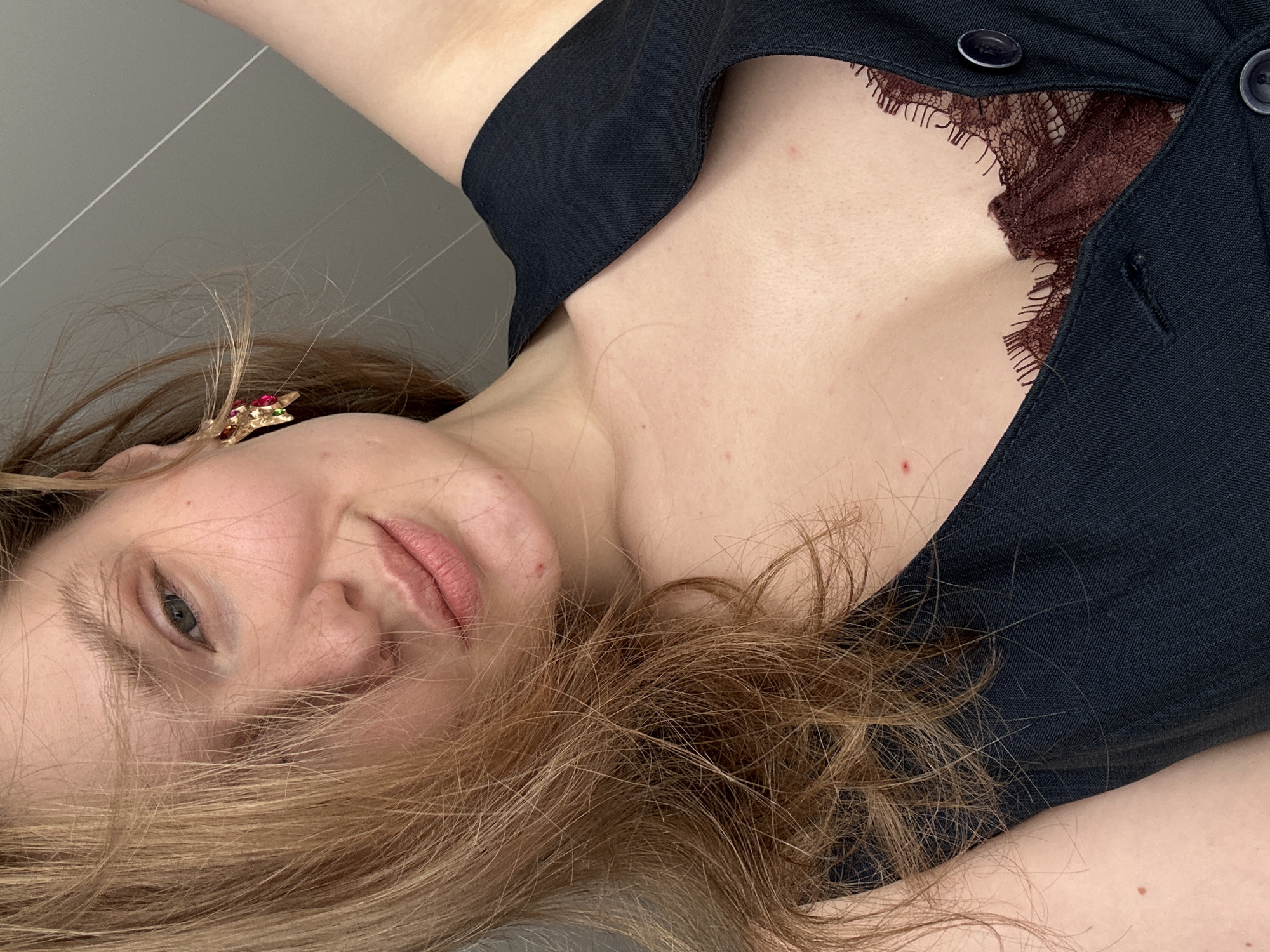 The image is a close-up, rotated view of a woman with fair skin and light brown hair. She is lying down, and her face is partially obscured by strands of hair. She has a single earring with red and green stones. She is wearing a dark-colored garment, likely a button-up top, over a dark red lace top, partially visible at the neckline. The background is a plain light gray surface. There is no evidence to suggest the image was taken in Kazan, Russia.