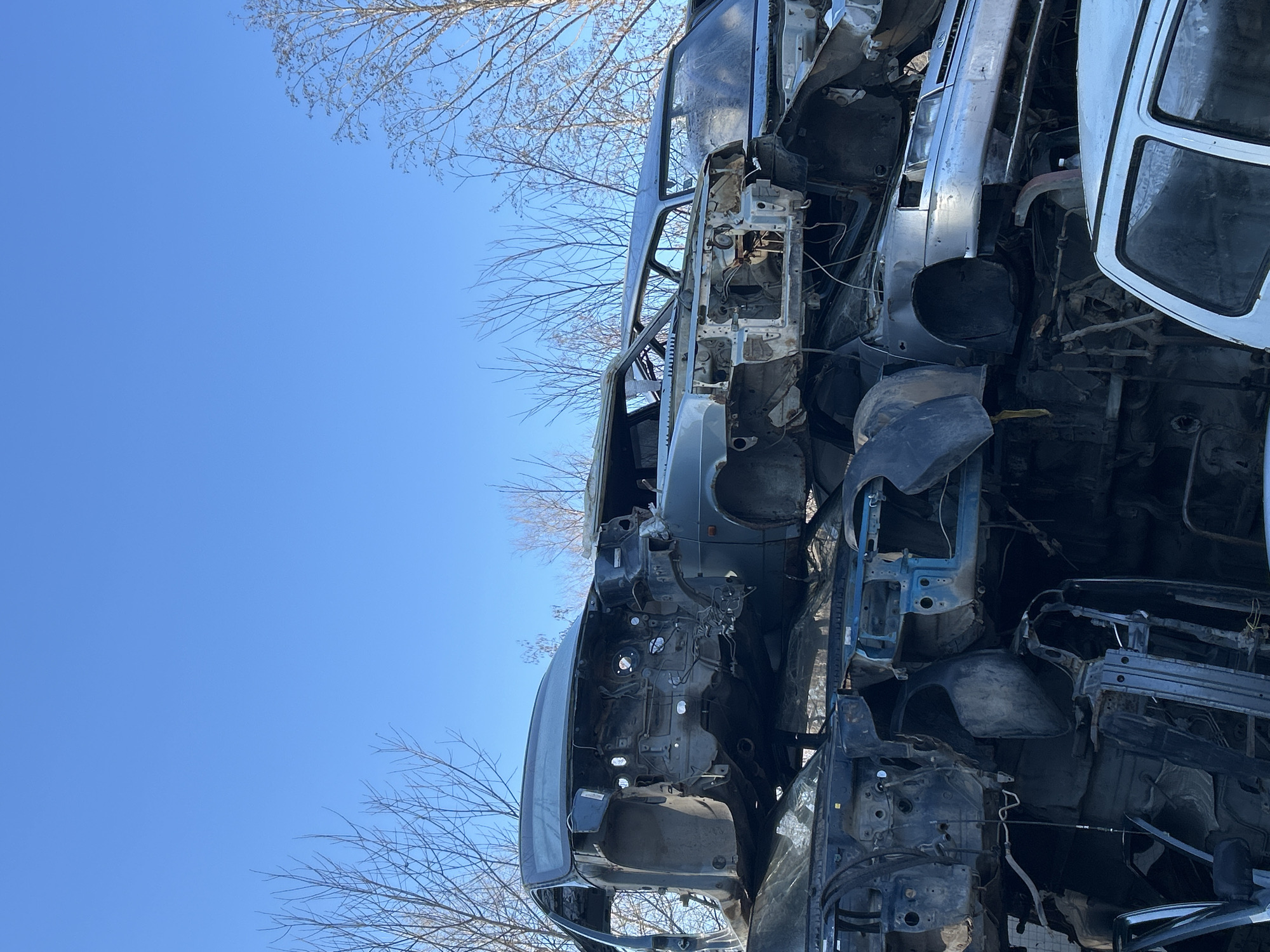 The image shows a stack of mangled car frames and chassis in an outdoor setting. The metal is heavily damaged and rusted. A clear blue sky and bare tree branches are visible in the background. Without additional information, it is not possible to confirm the location is Chaikovsky, Russia. There are no people visible in the image.