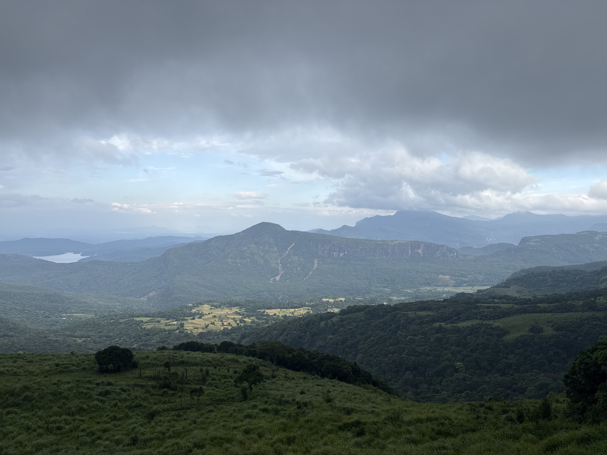 The image presents a landscape view from Illukkumbura, Sri Lanka, under a cloudy sky. The foreground is characterized by a grassy hill, dotted with small trees and bushes. The midground features a valley with dense forests and agricultural lands, with a prominent mountain in the center of the frame. Further back, a lake is visible amidst the layers of hills and mountains. The sky is covered with heavy clouds, suggesting overcast weather conditions. There are no people, subjects, or actions depicted in the view.