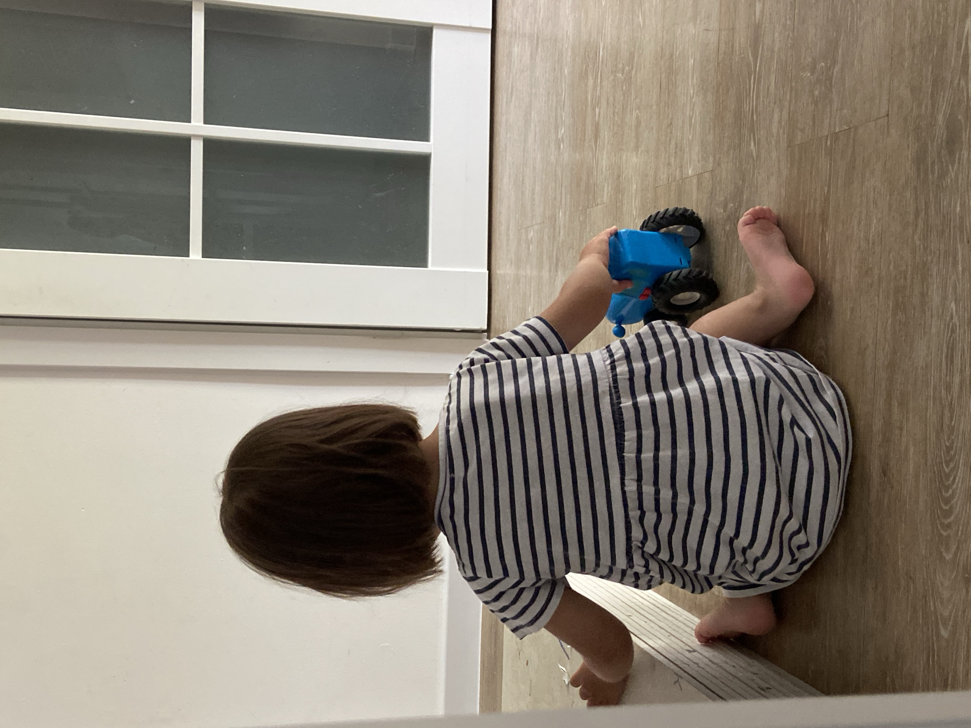This overhead, slightly angled shot captures a young child playing indoors. The child, seen from behind and likely a toddler, is positioned on a light brown, wood-patterned floor. They are wearing a white garment adorned with vertical navy blue stripes and have short, dark brown hair. The child appears to be on their hands and knees or kneeling, actively engaging with a blue toy tractor that features black wheels. Their bare feet are visible, one foot tucked under them and the other partially extending to the side.
To the left of the child, a prominent white architectural feature occupies a significant portion of the frame. This structure, possibly an interior door or a large window panel, is characterized by white framing and mullions that create a grid pattern. The glass within these frames appears frosted or opaque, obscuring any view beyond it. The overall lighting is bright and even, consistent with an indoor setting during the daytime. No specific location cues, weather conditions, or explicit emotions are discernible from the image, and there is no visible text.