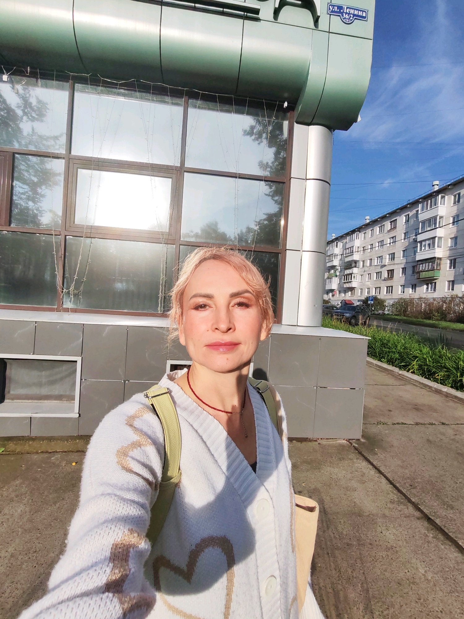 A blonde woman with pink-toned hair is taking a selfie outdoors on a sidewalk in front of a modern building with large windows. She is wearing a light blue cardigan with a heart pattern and a green strap over her shoulder. A red necklace is visible around her neck. Behind her, a multi-story apartment building and a street with parked cars are visible. The sky is blue with some scattered clouds. A street sign with Russian lettering, "ул. Ленина 36/2", is affixed to the building. The sun is shining, casting shadows on the ground.
