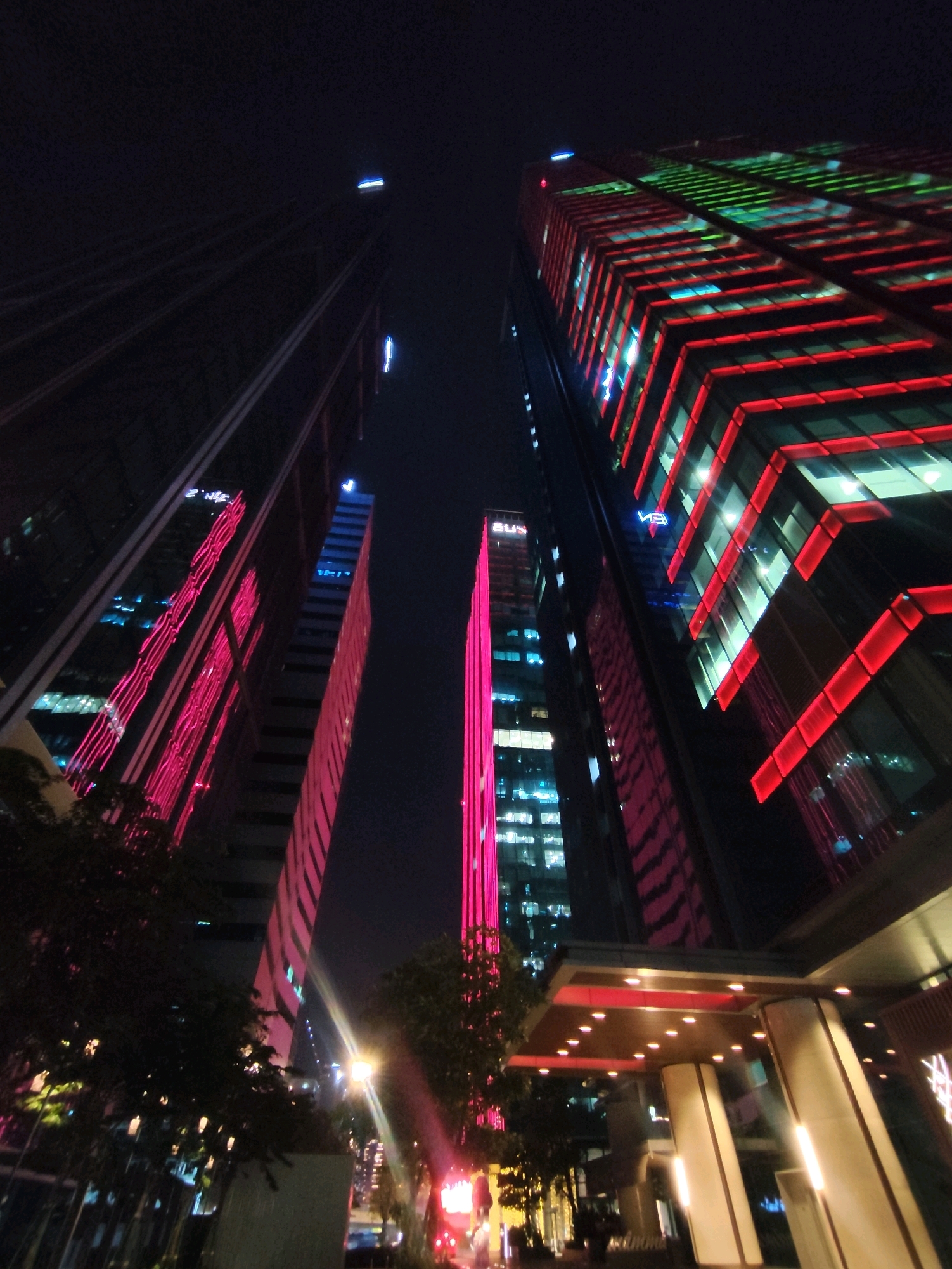 This is a low-angle, wide shot of skyscrapers at night in Ho Chi Minh City, Vietnam. The buildings are illuminated with vibrant neon lights, primarily in shades of pink, red, and green. The skyscrapers are tall and modern, with glass facades that reflect the city lights. In the foreground, there is a dark silhouette of trees and streetlights. The sky is dark, indicating it is nighttime. The image captures the dynamic and colorful urban landscape of Ho Chi Minh City after dark.