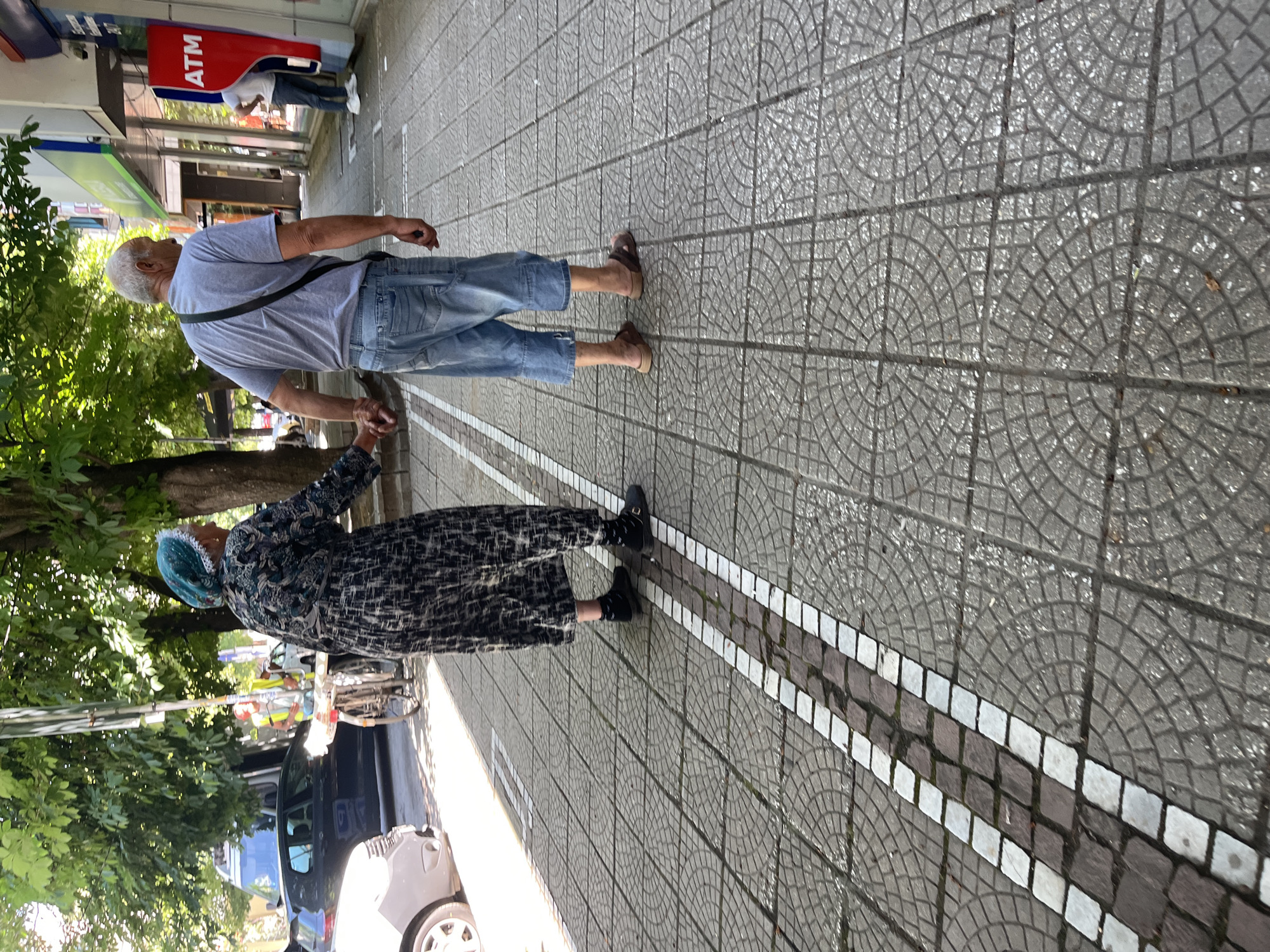 The image shows an elderly couple walking on a tiled sidewalk in Pazardzhik, Bulgaria, holding hands. The woman, on the left, wears a patterned dress and a blue headscarf. The man, on the right, wears a grey t-shirt, denim shorts, and sandals. They are walking away from the camera towards an ATM machine visible on the right side of the image. The sidewalk is paved with grey square tiles featuring a circular pattern. A narrow strip of alternating white and dark square tiles runs along the left side of the sidewalk. In the background, trees provide shade, and a street with parked cars and a person near a bicycle is visible.