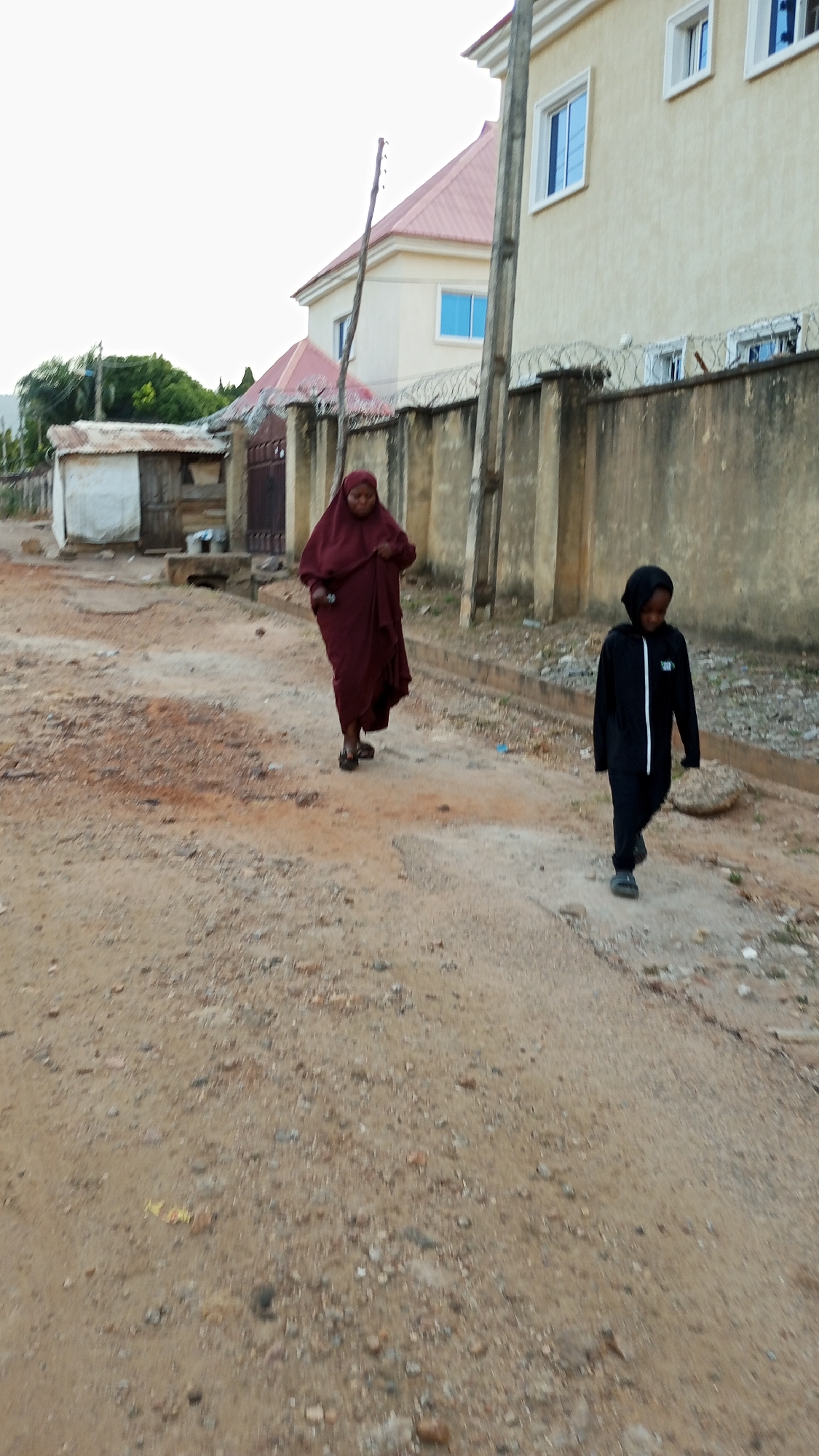 A woman wearing a dark red hijab and matching robe walks on a dirt road in Adankolo, Nigeria. To her right is a young person in a black hooded tracksuit. The road is unpaved with loose dirt and small rocks. A concrete wall topped with barbed wire borders the road on the right. Behind the wall is a two-story beige building with a red roof. Across the road is a small, dilapidated structure. Overhead power lines are visible.