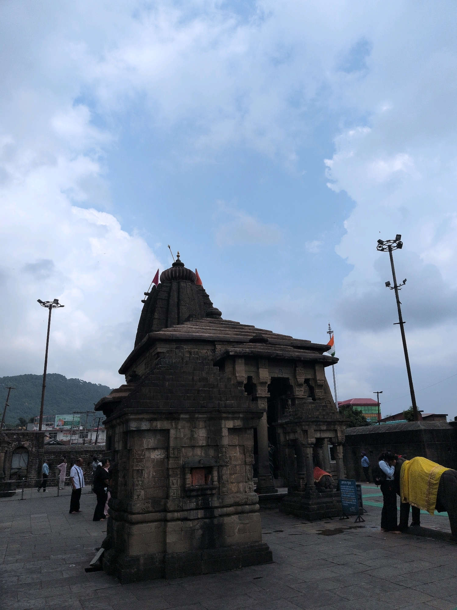 An overcast sky with dramatic clouds looms over the ancient stone temple of Baijnath Mahadev in Baijnath, India. The temple, constructed from dark, weathered stone, features intricate carvings and a multi-tiered roof adorned with red flags. Two tall light poles with multiple lamps stand to the left and right of the temple. In the foreground, a stone-paved courtyard is visible, with several people walking around. To the right, an elephant, draped in a bright yellow cloth, stands with its head lowered, possibly interacting with a person. The temple complex appears to be an active religious site, with devotees and possibly tourists present. In the background, a lush green mountain range is visible, hinting at the temple's scenic location. Buildings and wires are also visible in the distance, indicating the presence of a town or city.