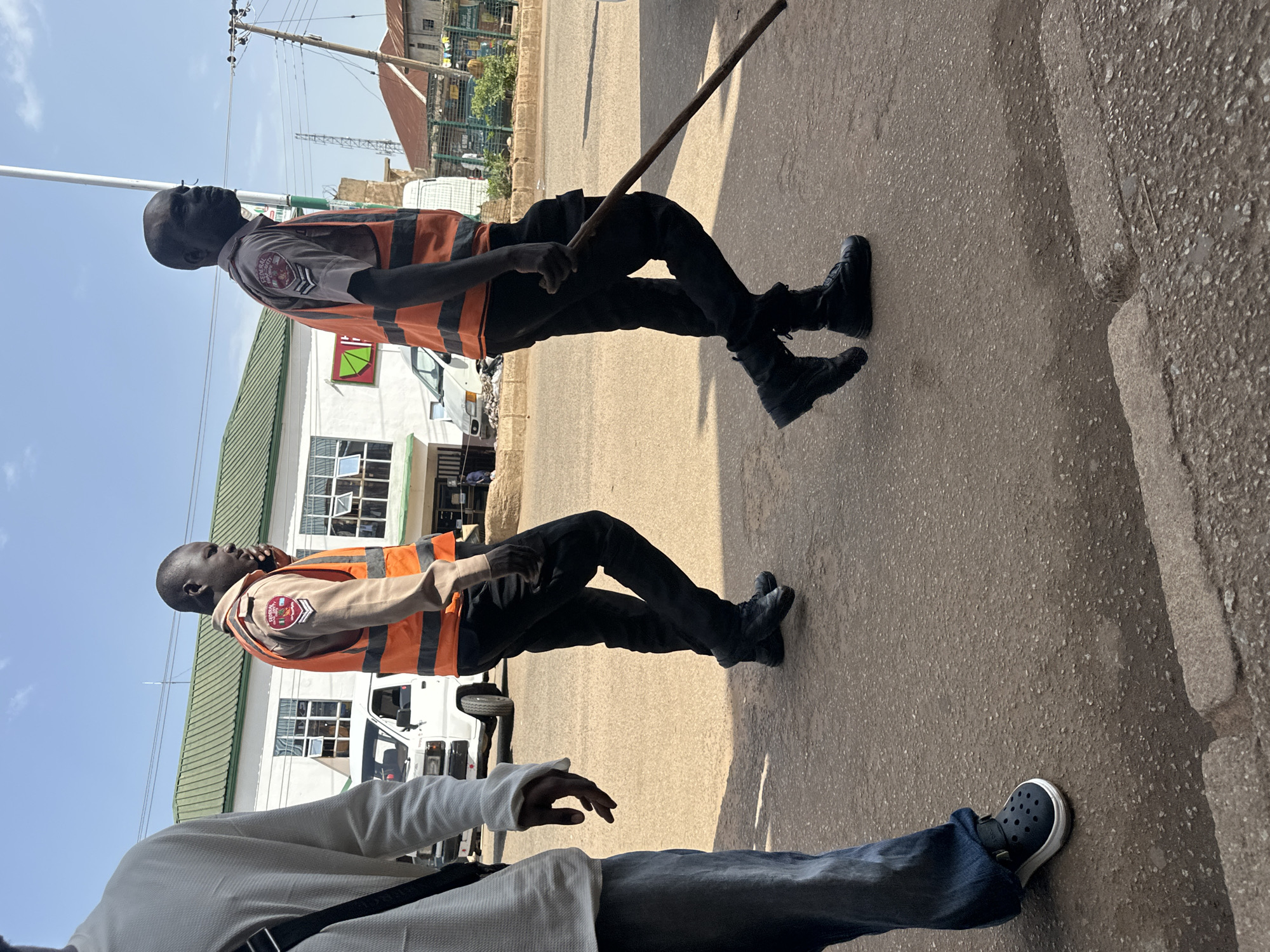 In Bukuru, Nigeria, three individuals are walking along a paved road under a clear sky. The person on the left, partially visible and facing away, wears a white long-sleeved shirt and blue trousers. The two individuals in the center and right are wearing orange safety vests over collared shirts and black trousers. Both are also wearing black boots. The person in the center, facing slightly left, is holding a mobile phone to their ear. The person on the right, facing forward, is carrying a long wooden stick. Behind them, a white van is parked on the side of the road, and buildings with green roofs line the street. Electrical poles and wires are visible against the sky, indicating an urban or semi-urban environment. The ground is a mix of asphalt and rougher concrete or stone.