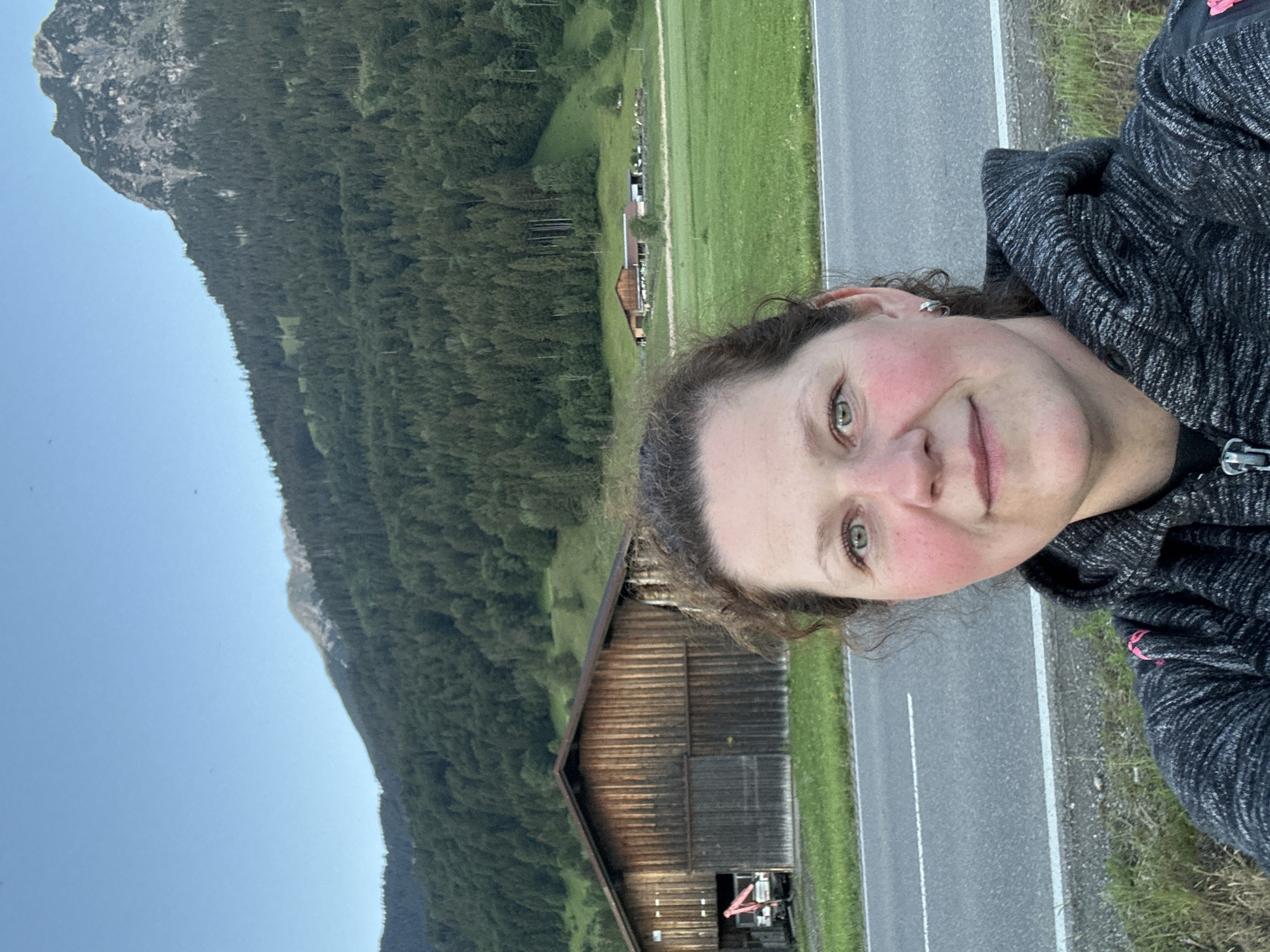 A woman is captured in a selfie against a picturesque mountainous landscape, likely in Oberstdorf, Germany. She occupies the right side of the frame, looking towards the viewer with a slight, closed-mouth smile. Her face shows rosy cheeks, green eyes, and she has dark, wavy hair, with a small silver earring visible on her left ear. She is wearing a grey and black patterned hoodie or jacket.
Behind her, a paved road with a solid white line and a dashed white line in the center stretches across the mid-ground. Beyond the road, vibrant green fields extend into the distance, dotted with several traditional wooden buildings, including a prominent barn-like structure with a dark wooden facade and a gabled roof. The background is dominated by a majestic mountain range, densely covered with dark green evergreen forests. Rocky outcrops are visible near the peaks on the far left side of the mountains. The sky is a clear, bright blue, indicating fair weather. The lighting is soft and even, suggesting either early morning or late afternoon. The scene conveys a tranquil, natural setting characteristic of an Alpine or pre-Alpine region. No visible text is present.