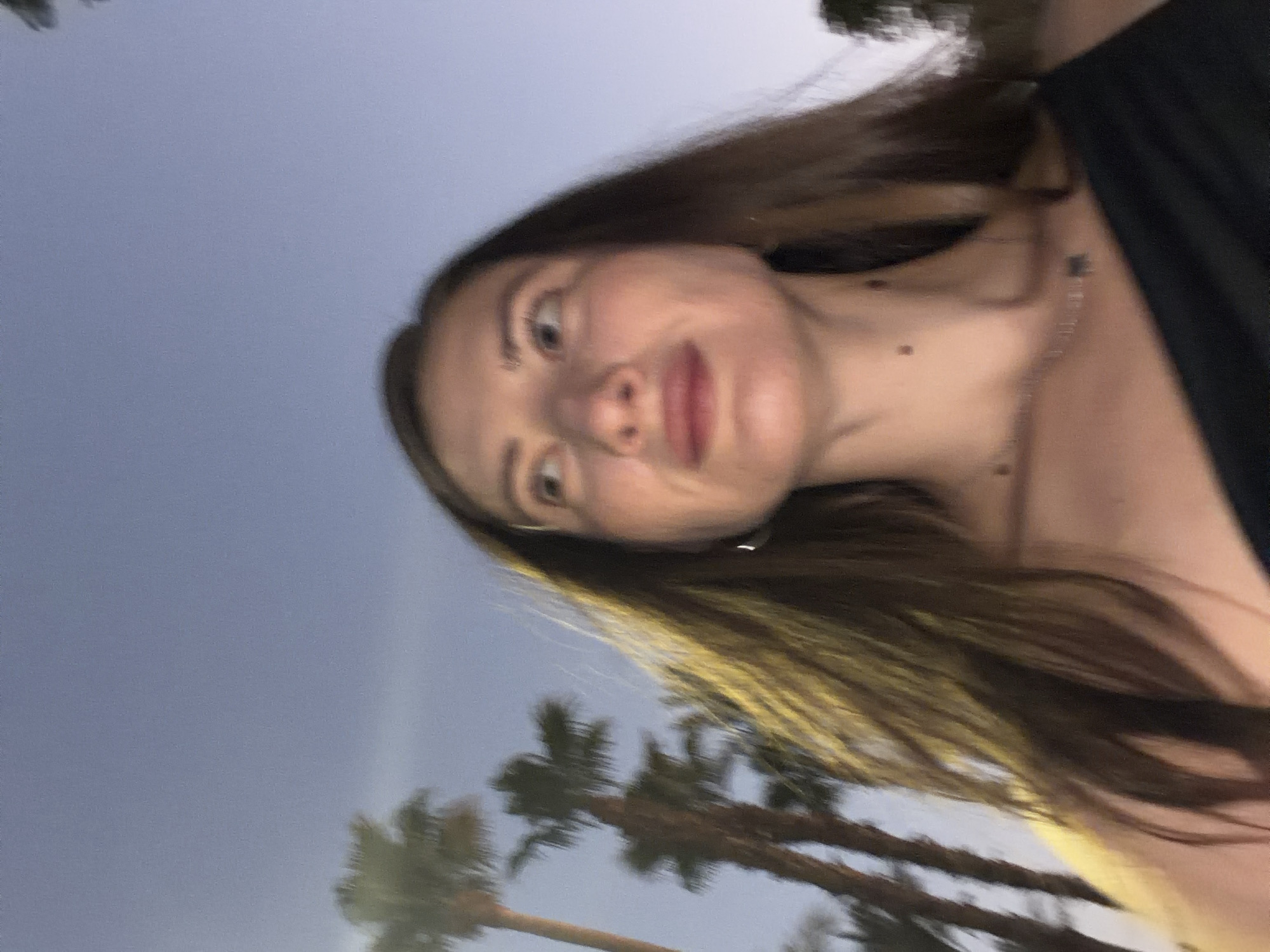 The image is a blurry, low-resolution, outdoor selfie taken at dusk. A woman with long, dark hair is in the foreground, her face turned to her left, looking upwards. She is wearing a black off-the-shoulder top and a delicate silver necklace. Palm trees are visible in the background, suggesting a tropical or subtropical location. The sky is a pale blue, characteristic of twilight. The overall impression is of a candid, informal moment captured in a warm climate. It is not possible to determine the specific location within Sharm El Sheikh, Egypt, from the provided image.