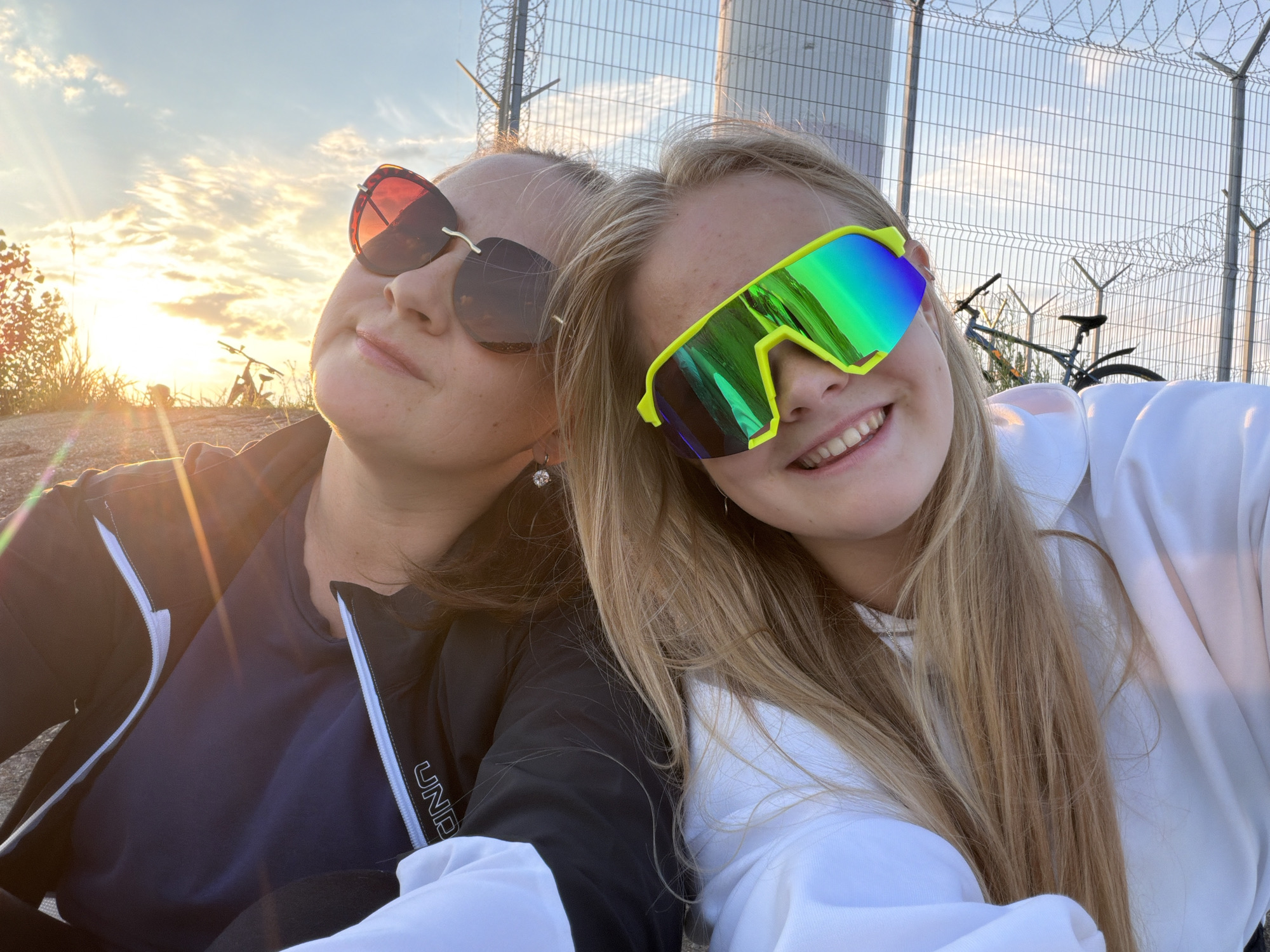 Two individuals, appearing to be young women, are captured in a close-up selfie outdoors. The person on the left, partially obscured by a black jacket and a navy blue shirt, wears large, red-tinted sunglasses. The person on the right, wearing a white hooded sweatshirt, sports oversized, neon yellow-framed sunglasses with reflective, multi-colored lenses. Both individuals have blonde hair. The background features a chain-link fence with barbed wire, suggestive of a secured area, and a utility pole. A bicycle is visible behind the fence, and the setting sun casts a warm glow across the sky, creating lens flares and illuminating the scene with golden light. The location is identified as Chaikovsky, Russia.