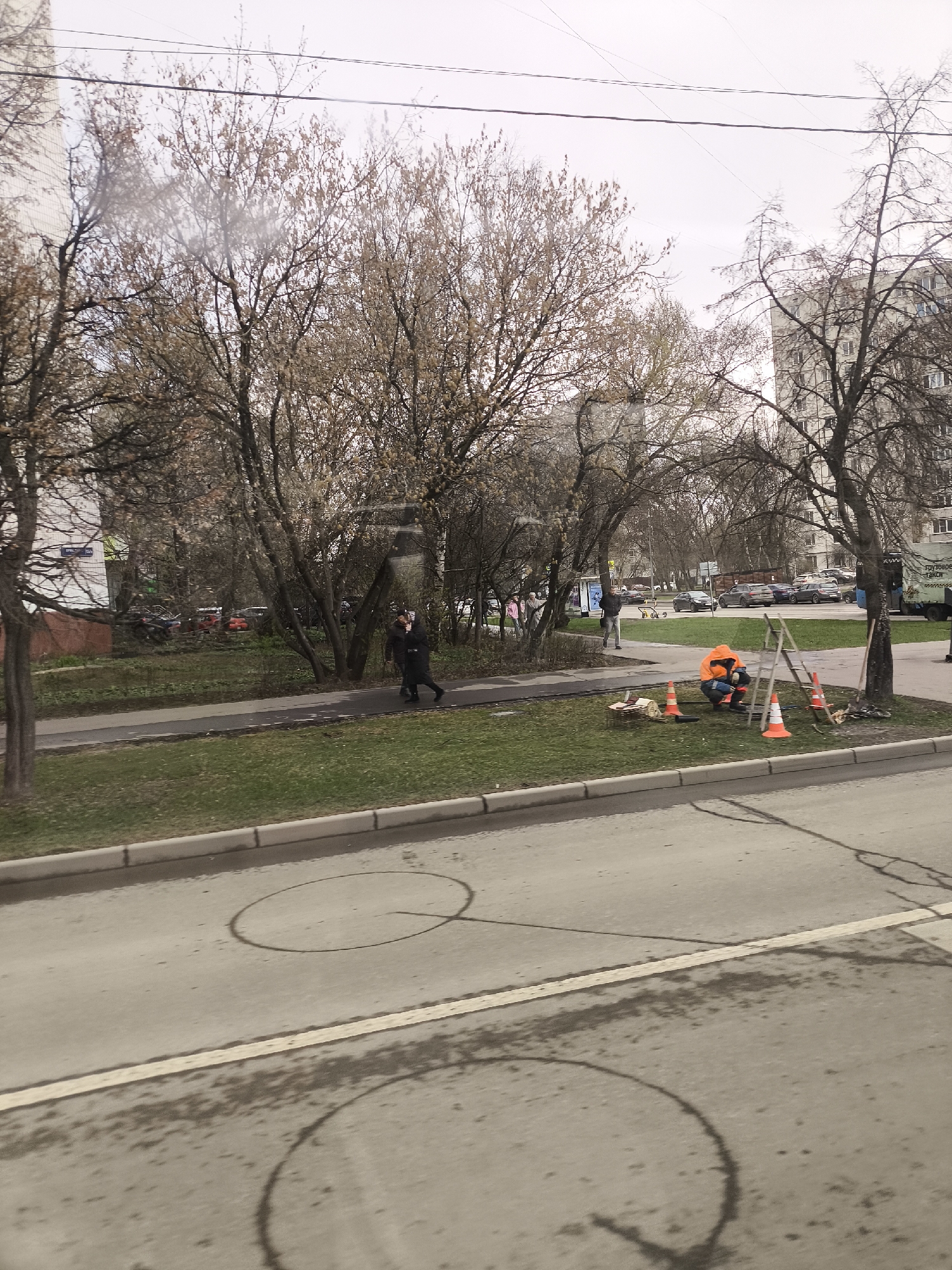 The scene depicts a street view in Moscow, Russia on a cloudy day. Several bare trees stand on either side of the street, with apartment buildings visible in the background. A man in a black coat walks on the sidewalk. A worker in an orange vest is kneeling on the grass next to a ladder and orange cones. The asphalt street in the foreground shows shadow reflections of overhead lighting or objects. Cars are parked along the street in the distance.