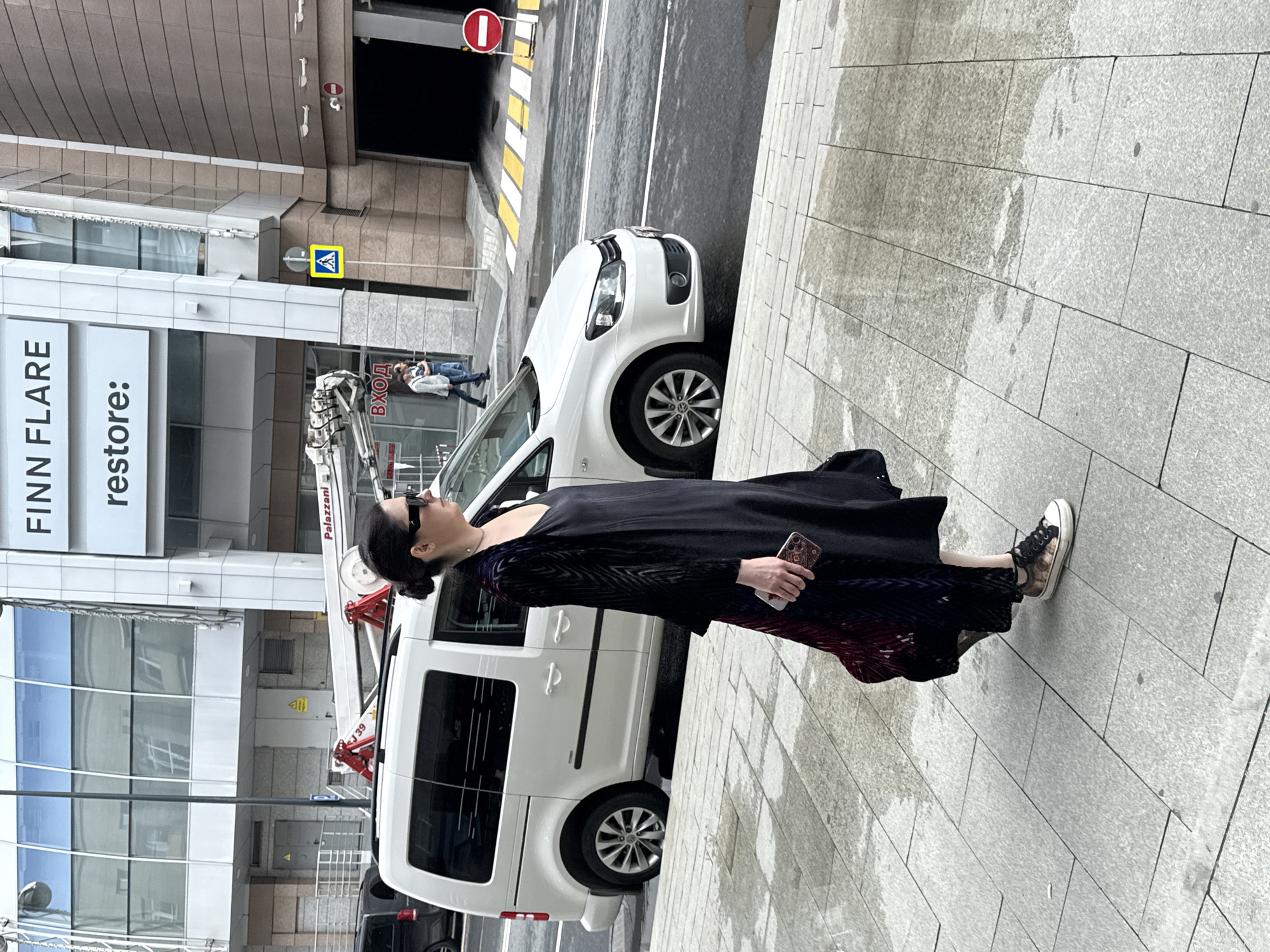 A woman with dark hair pulled back into a bun, wearing sunglasses, a long black dress, and a patterned velvet jacket, walks along a wet, tiled sidewalk. She is holding a smartphone in her right hand. A white van is parked on the street behind her, and to the right, a pedestrian crossing with yellow and white stripes is visible, alongside a parking garage entrance. The building behind the van has large glass windows and signage that reads "FINN FLARE" and "restore:". The scene appears to be in a commercial or urban area, possibly Moscow, Russia, given the presence of Cyrillic text on signage.