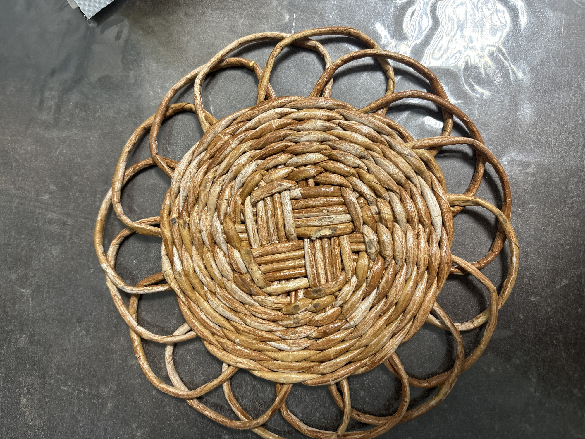 A close-up, overhead shot shows a round, woven placemat made of natural, light brown material. The placemat has a central woven square pattern, surrounded by concentric circles of woven material that extend outwards. These circles form an outer ring of decorative loops. The placemat rests on a dark, textured surface, possibly a countertop, with some light reflections and a small, white, crumpled object visible in the upper right corner. There are no people or actions depicted in the image. The location of Maidarovo, Russia is not discernible from the visual information provided.