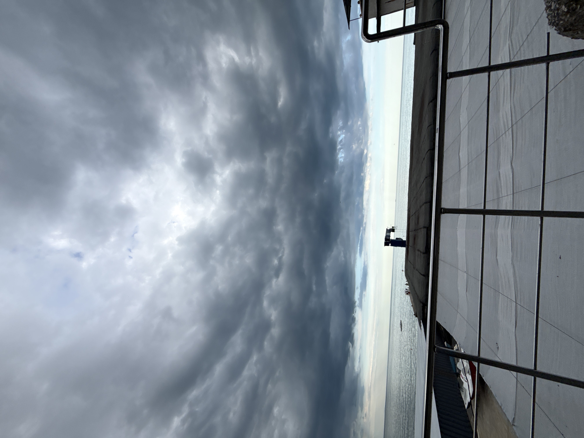 The image captures a dramatic, overcast sky filled with dark, swirling clouds. The sea is visible in the background, appearing calm with a muted, reflective surface. On the right side of the frame, a building's exterior is visible, featuring a white, slatted facade and a metal railing. The overall mood is somber and perhaps a little unsettling due to the intense cloud cover. The image appears to be taken during the daytime, though the heavy clouds obscure direct sunlight, creating a diffused and shadowed atmosphere. There is a faint glimpse of a distant structure or crane on the horizon above the sea. The perspective is from a slightly elevated vantage point, looking out towards the water and the sky.