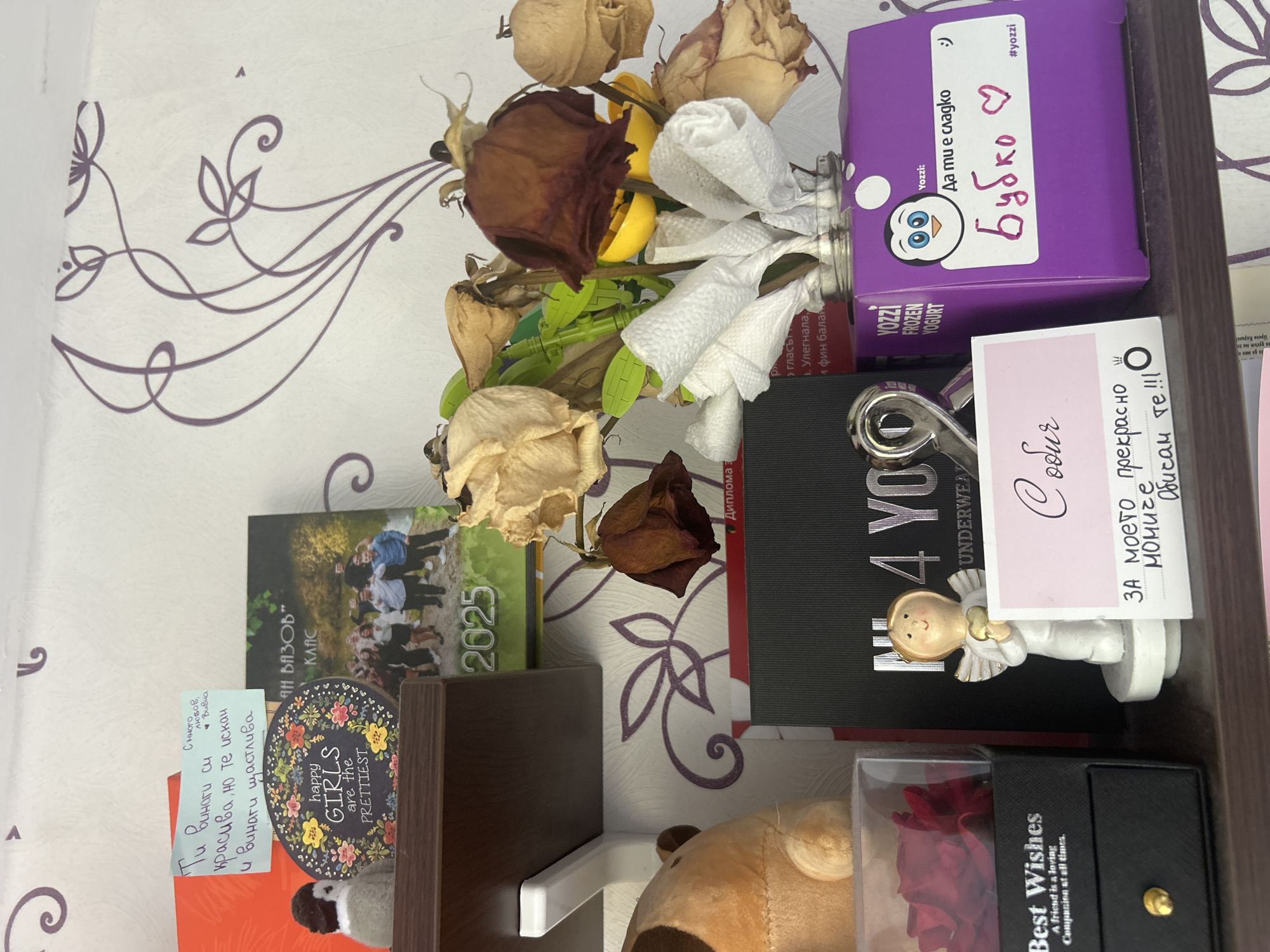 The image shows a shelf with various items on it. There are dried roses in a vase, a purple box of Yozzi frozen yogurt with a handwritten message "Бубко" (Bubko) and a heart, a photo album labeled "2025", a small sign saying "happy GIRLS are the PRETTIEST", a note with a handwritten message in Bulgarian, a box of underwear, and a small angel figurine. The background is a patterned wallpaper.