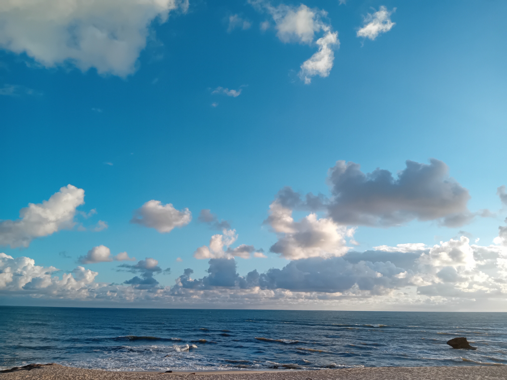 The image shows a beach scene with a calm sea under a partly cloudy sky. The sand in the foreground is light brown. The sea is a dark blue with small waves breaking on the shore. The sky is a bright blue with puffy white and gray clouds scattered throughout. A large rock is visible near the horizon. The location is unknown.