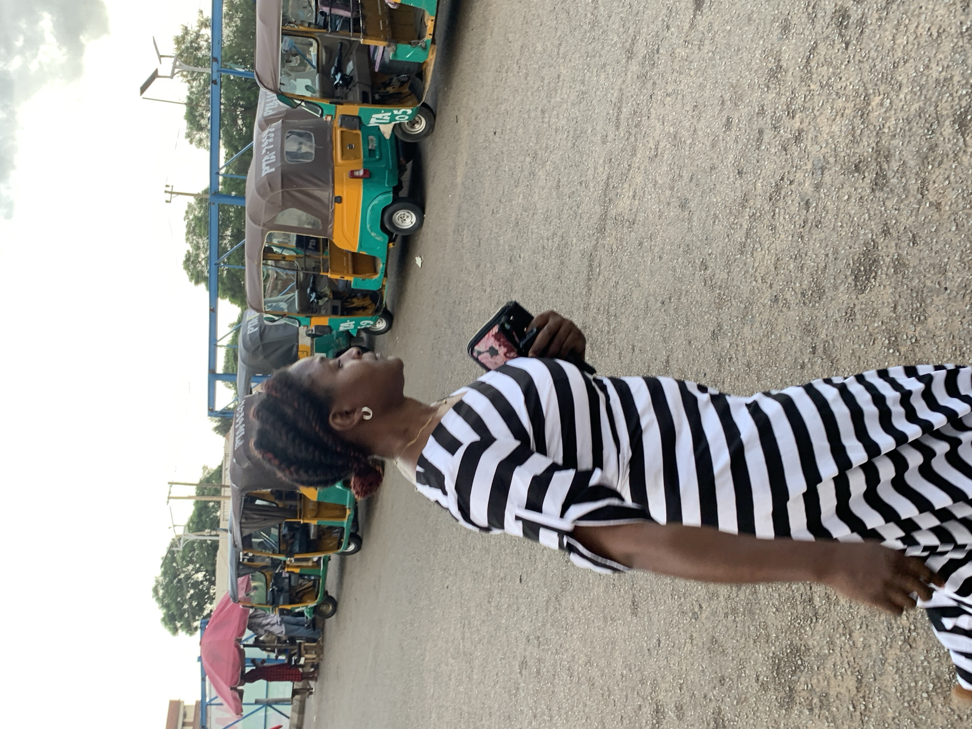 A woman wearing a black and white striped dress walks past a row of parked auto-rickshaws in Jos, Nigeria. The auto-rickshaws, also known as "Keke NAPEPs," are colorful, with green and yellow bodies and brown canopies. Some have license plates visible, such as "TA-7859" and "TA-705." In the background, a red umbrella shades a small market stall where several people are gathered. Trees and power lines are visible against a cloudy sky. The ground is paved with gravel. The woman is holding a dark-colored object in her right hand.