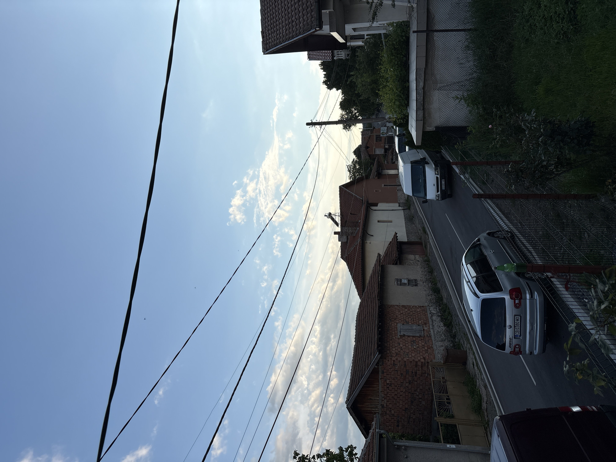 The image captures a street scene in Anton, Bulgaria, under a partly cloudy sky with a light blue hue. Multiple overhead power lines crisscross the frame, creating a sense of urban infrastructure. On the right side of the street, a modern white van is parked. Further down the road, a silver Volkswagen Sharan minivan is driving away from the camera, with the license plate "CB 0380 MX" visible. The street is lined with residential buildings, many featuring tiled roofs and brickwork. Some houses have visible solar panels on their roofs. Greenery, including trees and bushes, flanks the sides of the road and property boundaries. A metal fence runs along the right side of the street in the foreground. The overall impression is of a quiet, residential street in a small town.